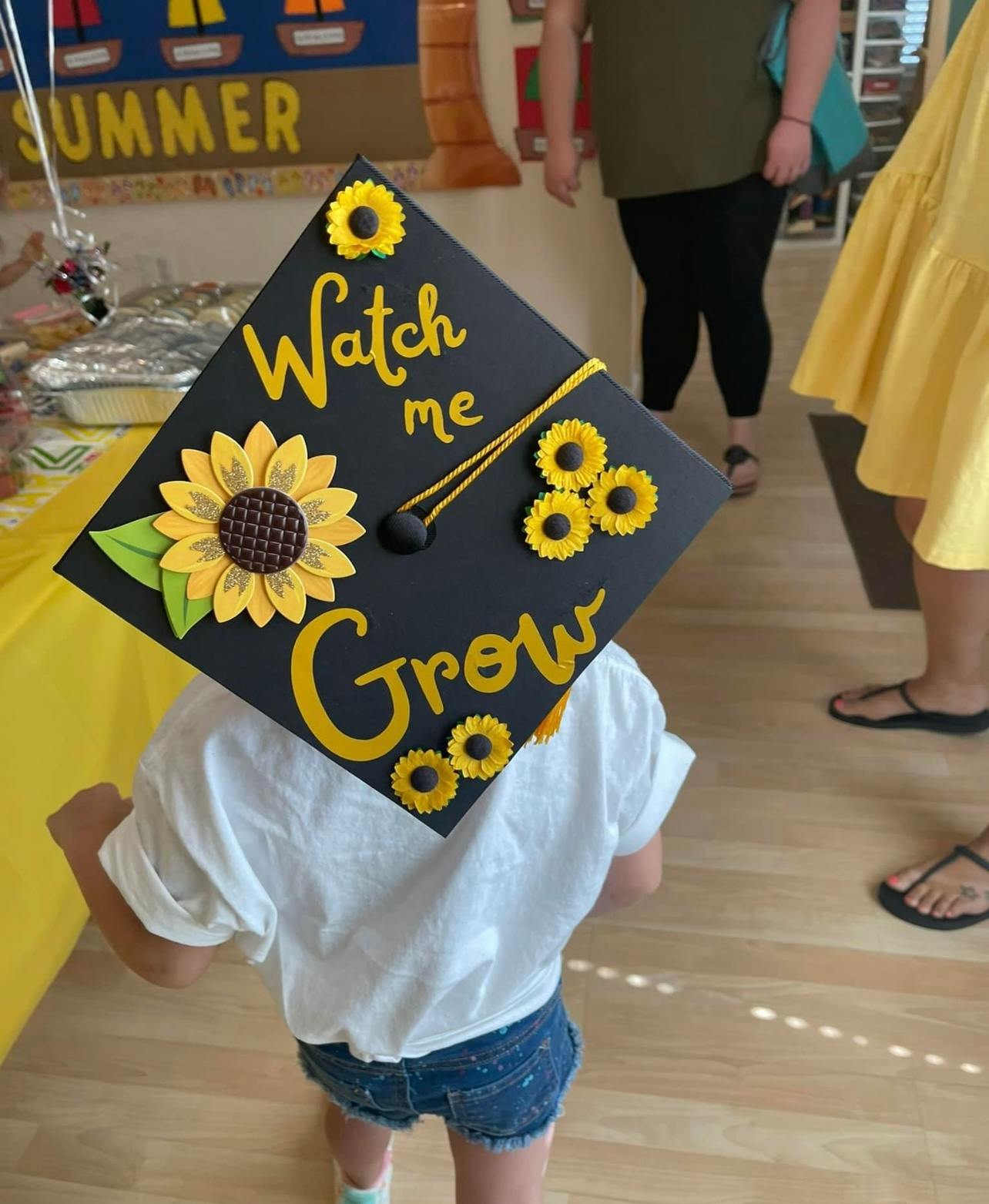 Child wearing a graduation cap decorated with sunflowers and "Watch me Grow"