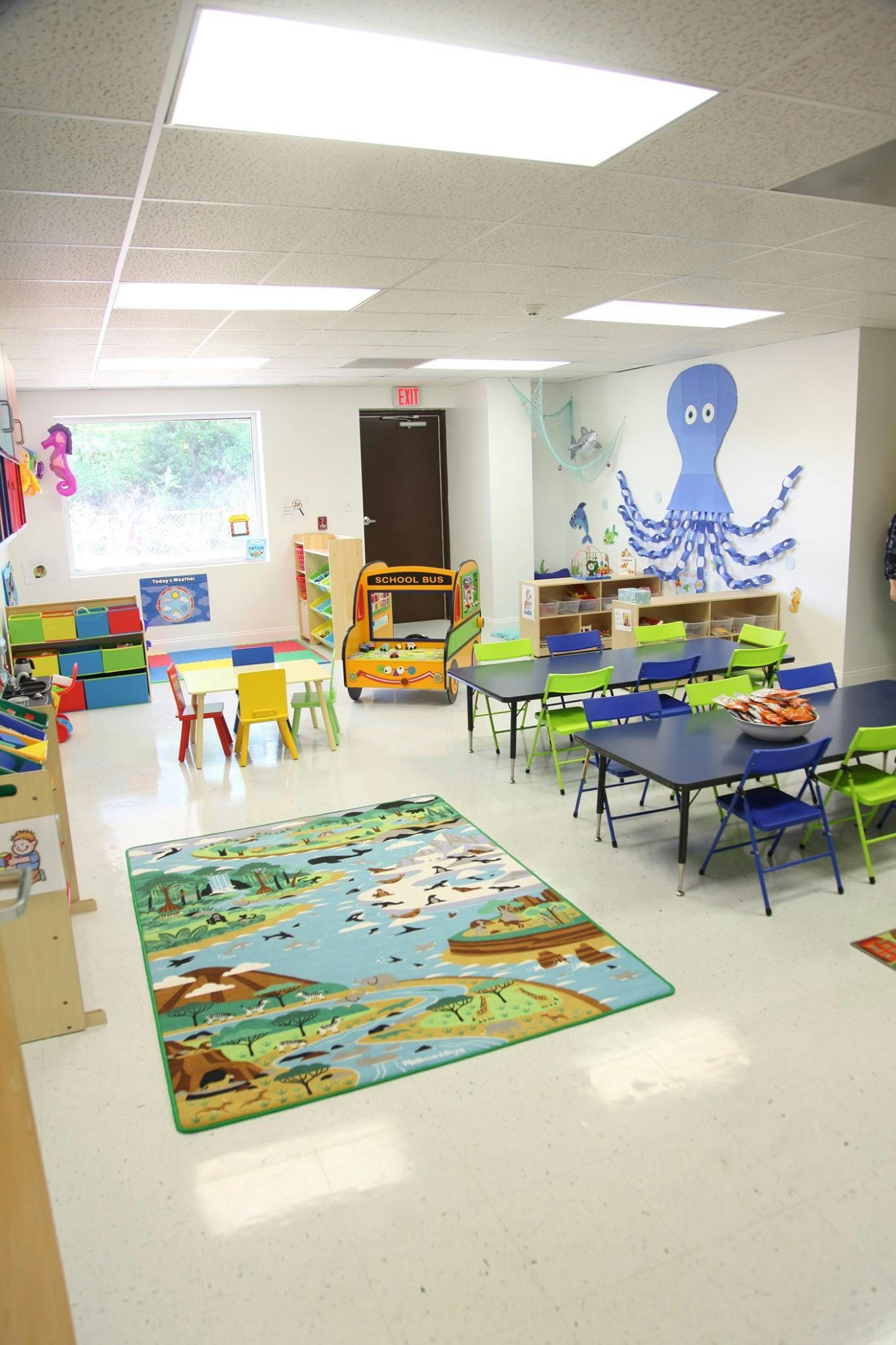 An empty classroom with small tables and chairs, a play school bus, and a colorful rug