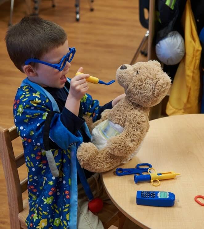 Child in glasses feeding a teddy bear with a toy spoon