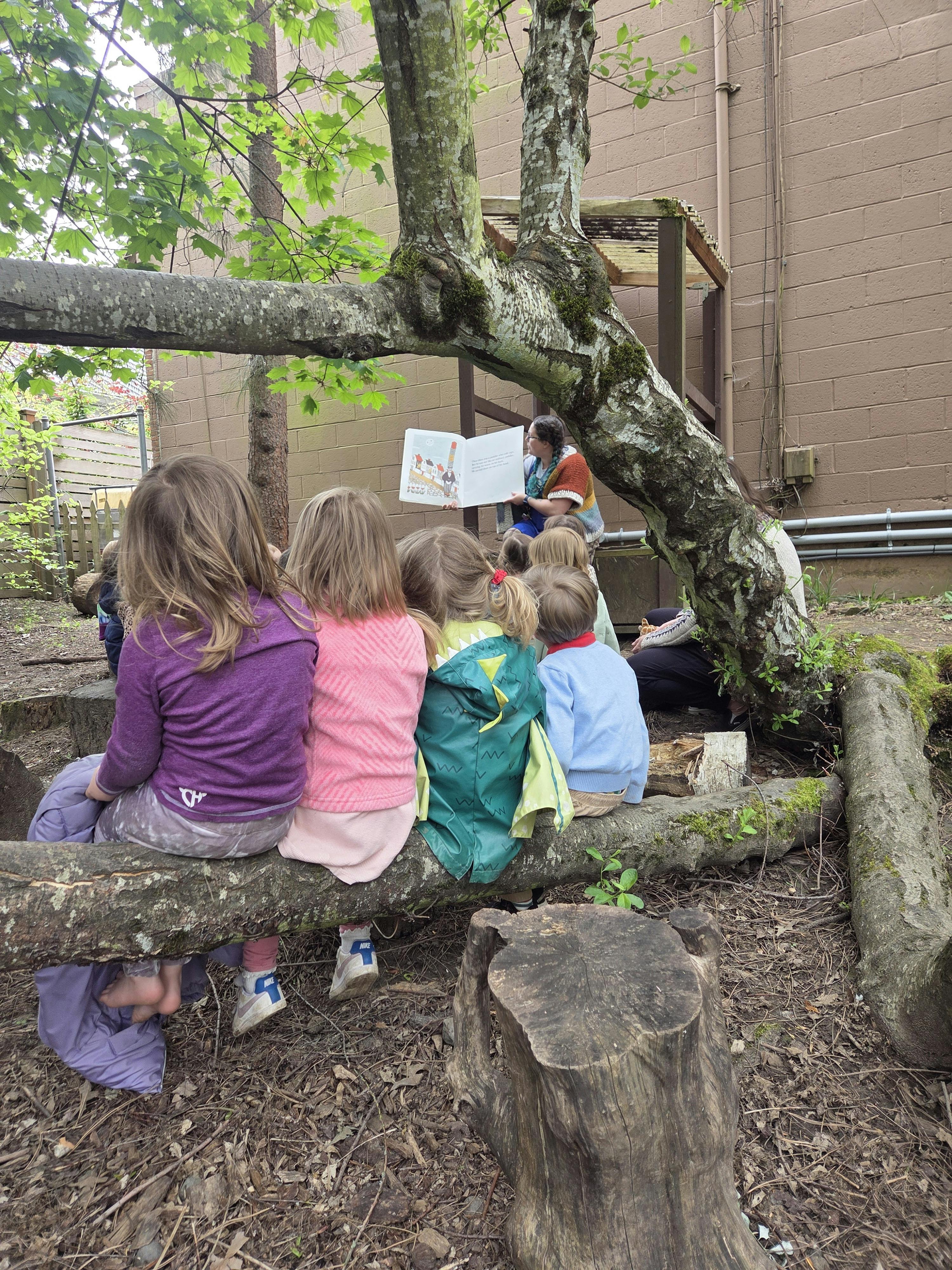 Children sitting on a log listening to a person read a book outdoors