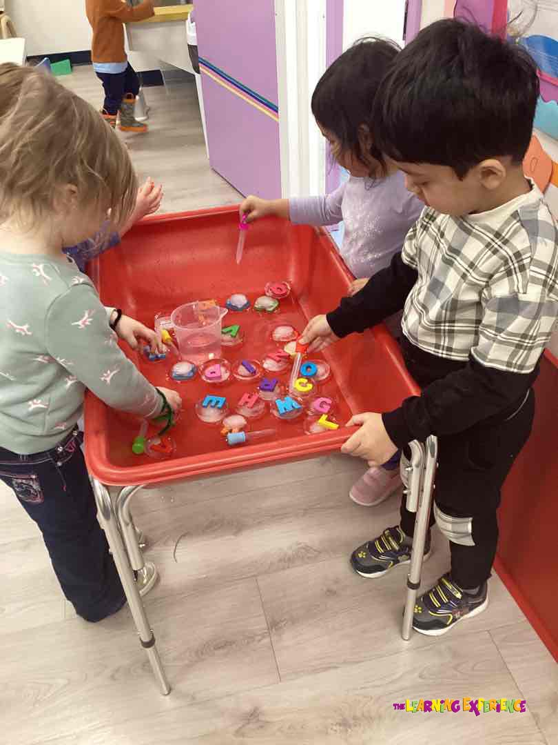 Three children playing at a red table with letters in ice