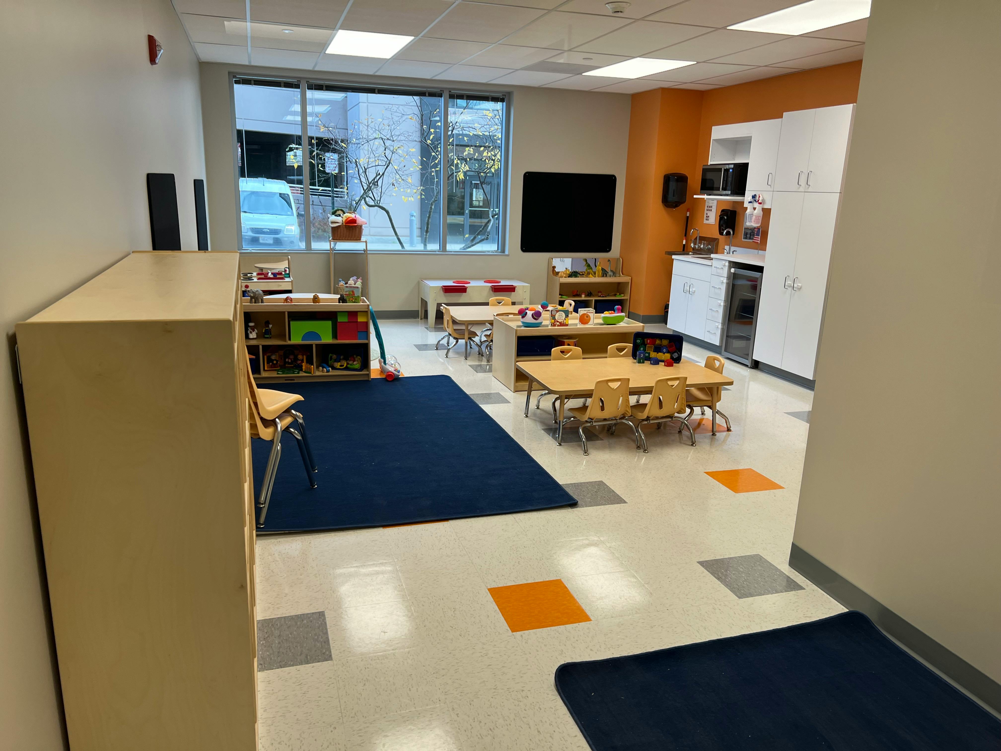 Classroom with small tables and chairs, a blue rug, and large windows