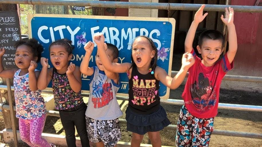 Five children with arms raised and mouths open in front of a "CITY FARMERS" sign