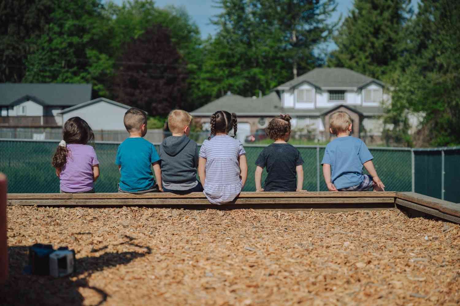 Children sitting on a wooden border at a playground, viewed from behind