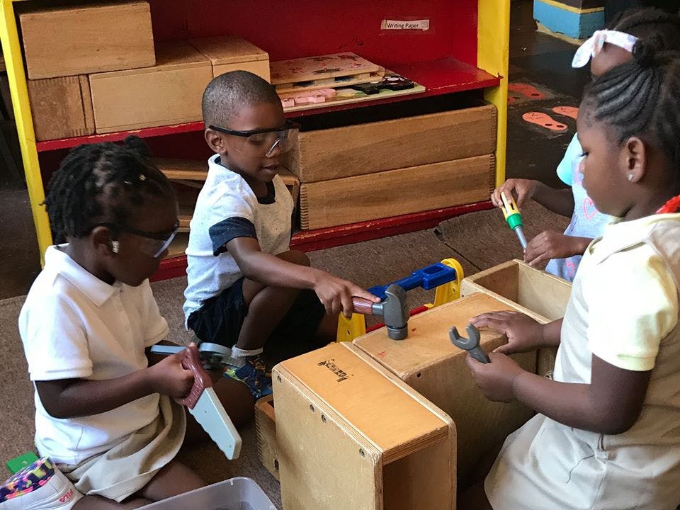 Three children playing with toy tools and wooden blocks in a classroom