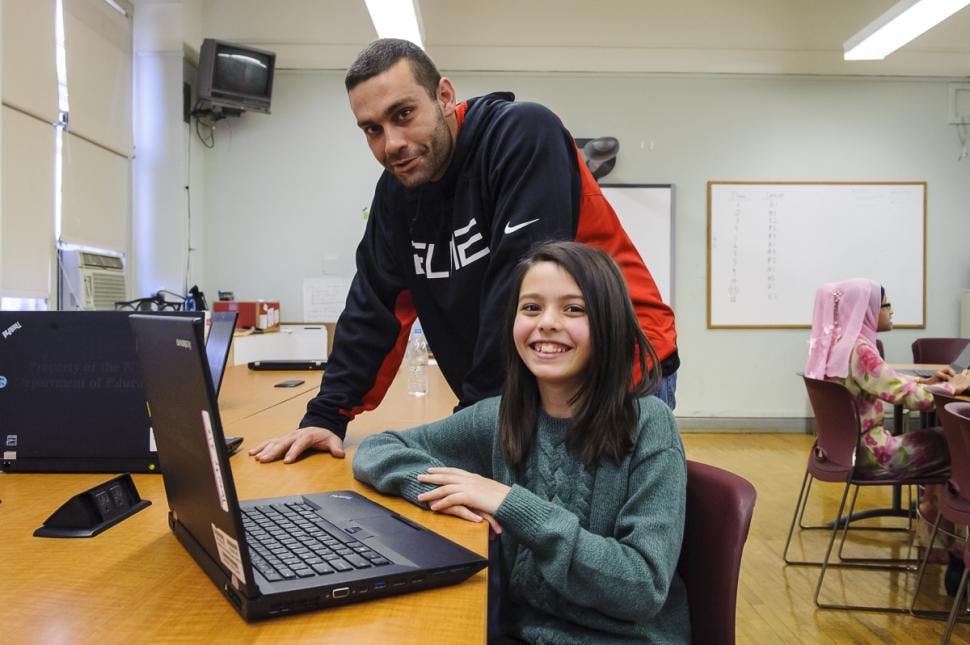 Person standing behind a child seated at a laptop in a classroom