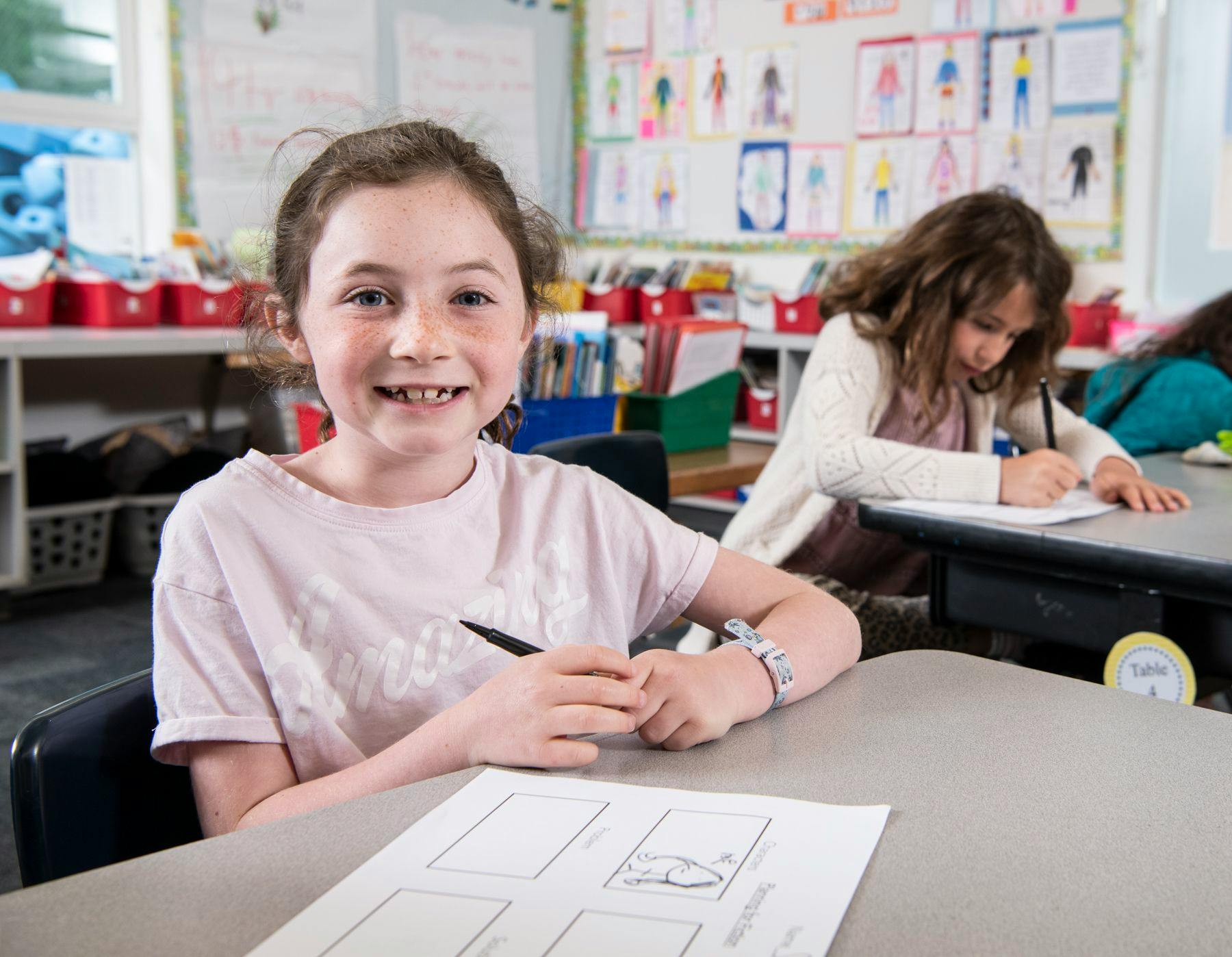 Child in a classroom holding a pen at a desk with another child drawing nearby