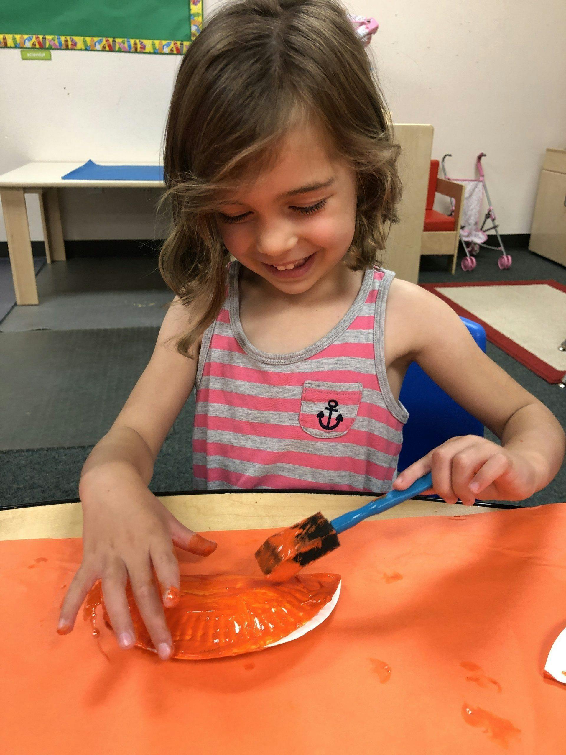 Child painting a paper plate orange with a brush in a classroom