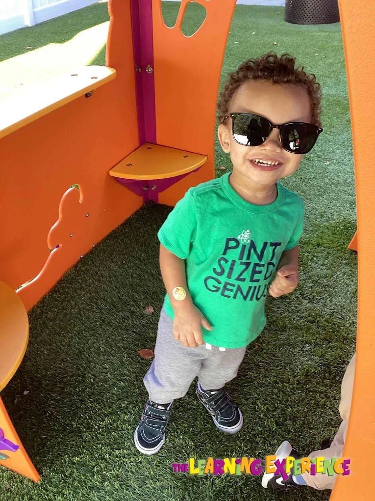 Child wearing sunglasses and a green shirt that says "Pint Sized Genius"
