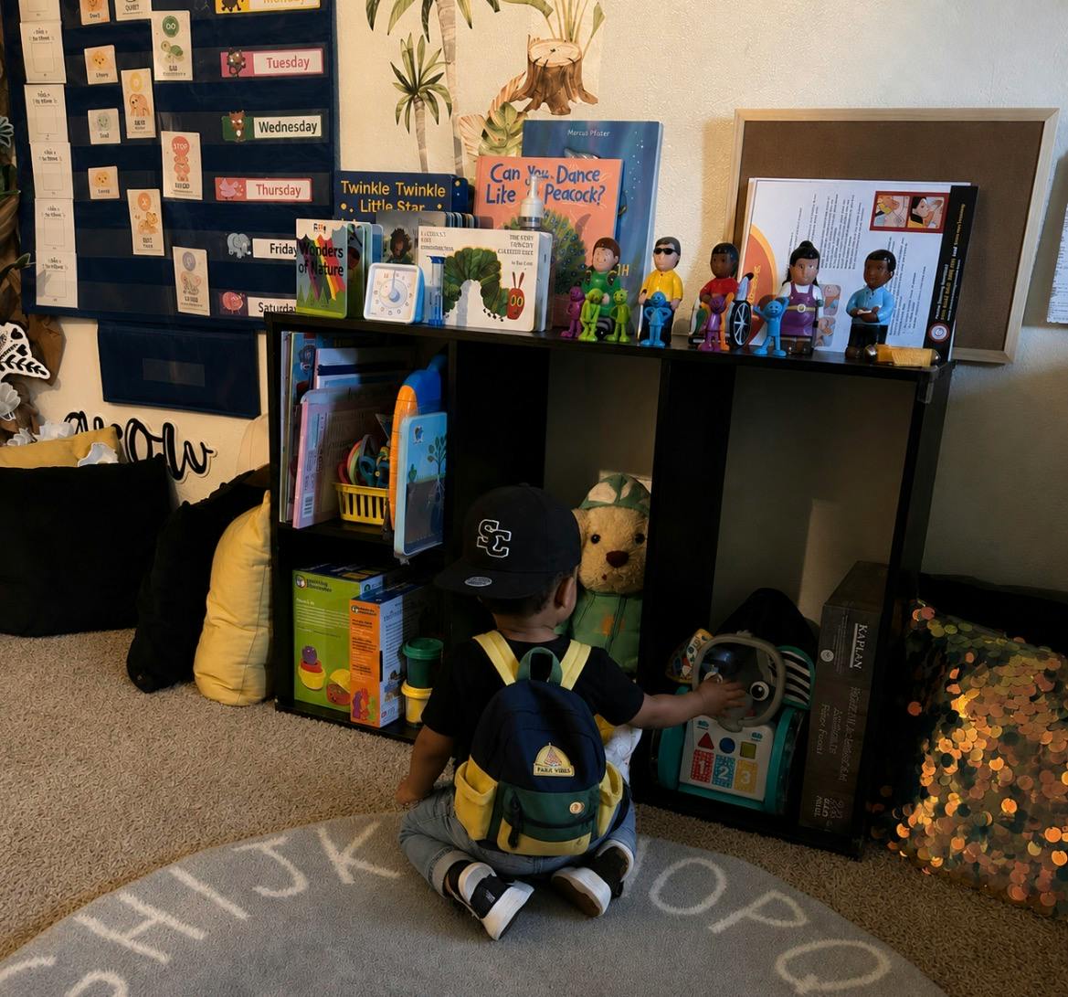 Child wearing a backpack and hat sitting on the floor by a toy shelf