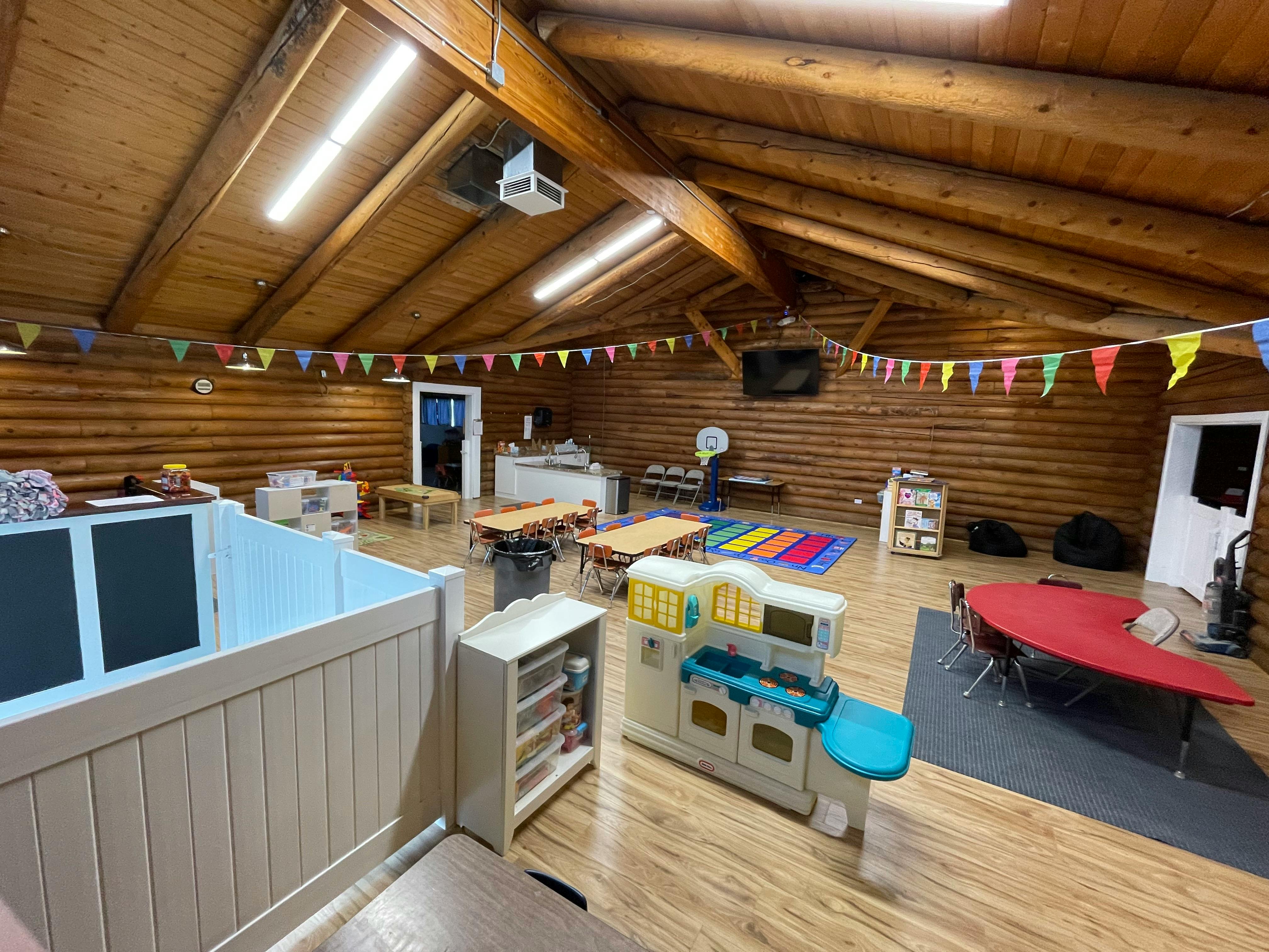 Indoor play area with wooden walls and ceiling, tables, and play kitchen