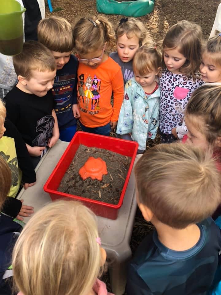 Children gathered around a red bin with dirt and an orange object