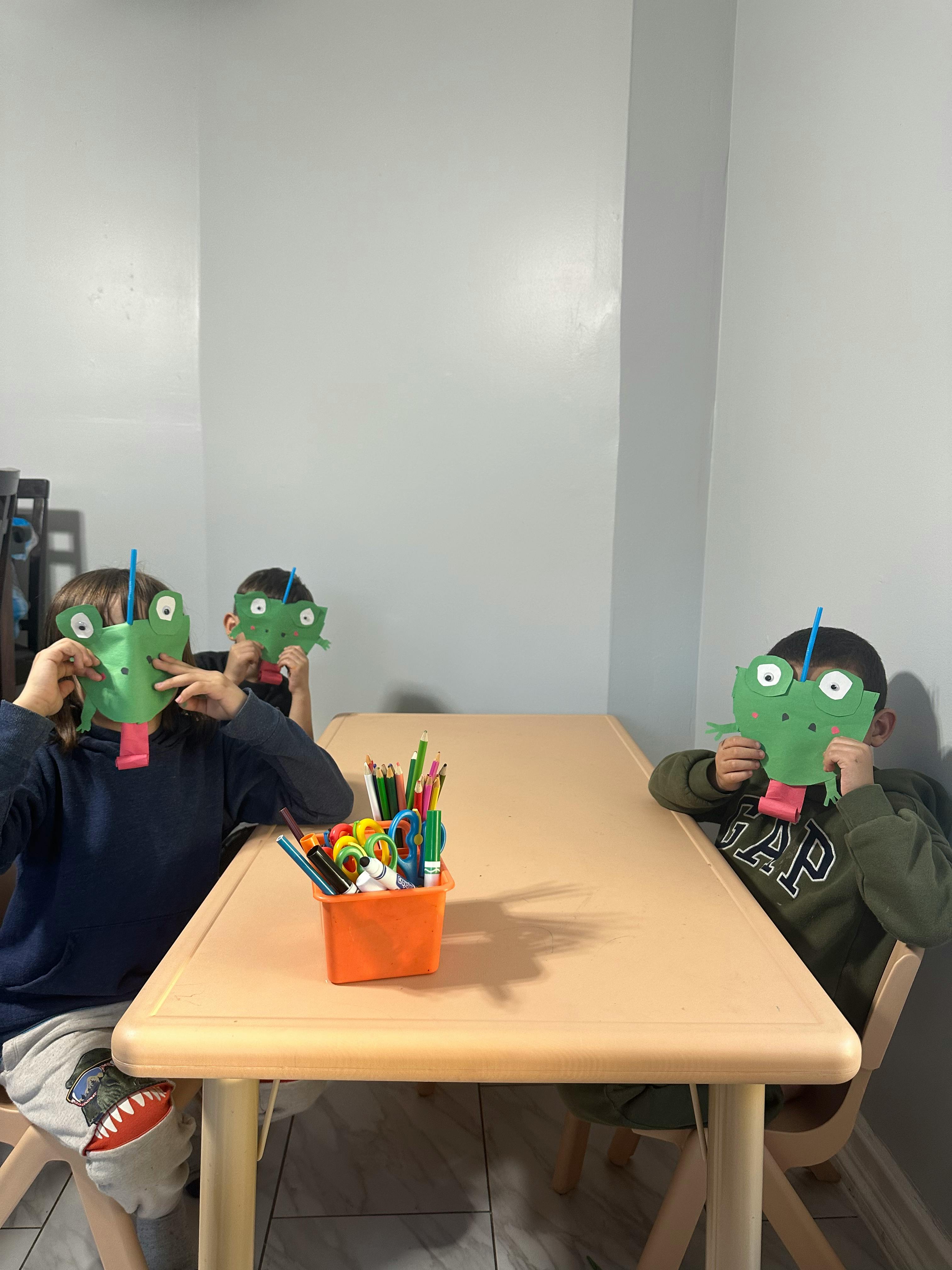 Three children wearing green paper masks at a table with art supplies