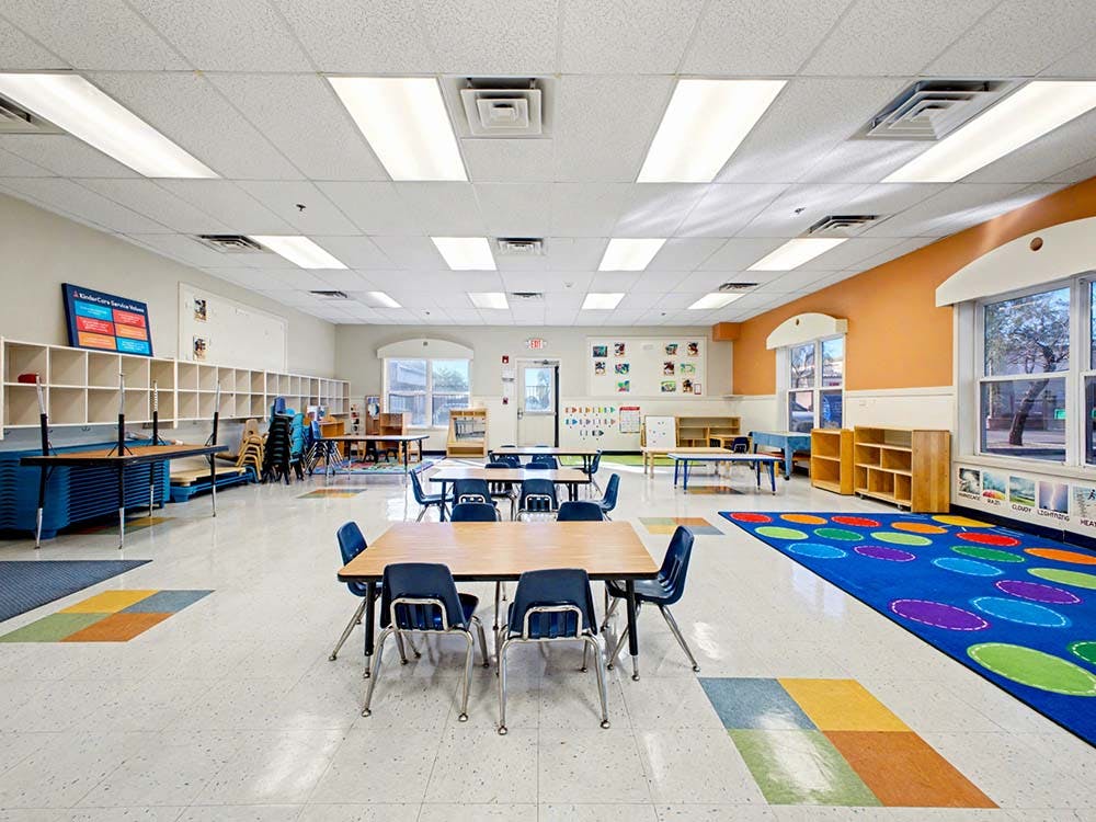 empty classroom with small tables and chairs, shelves, and a colorful rug