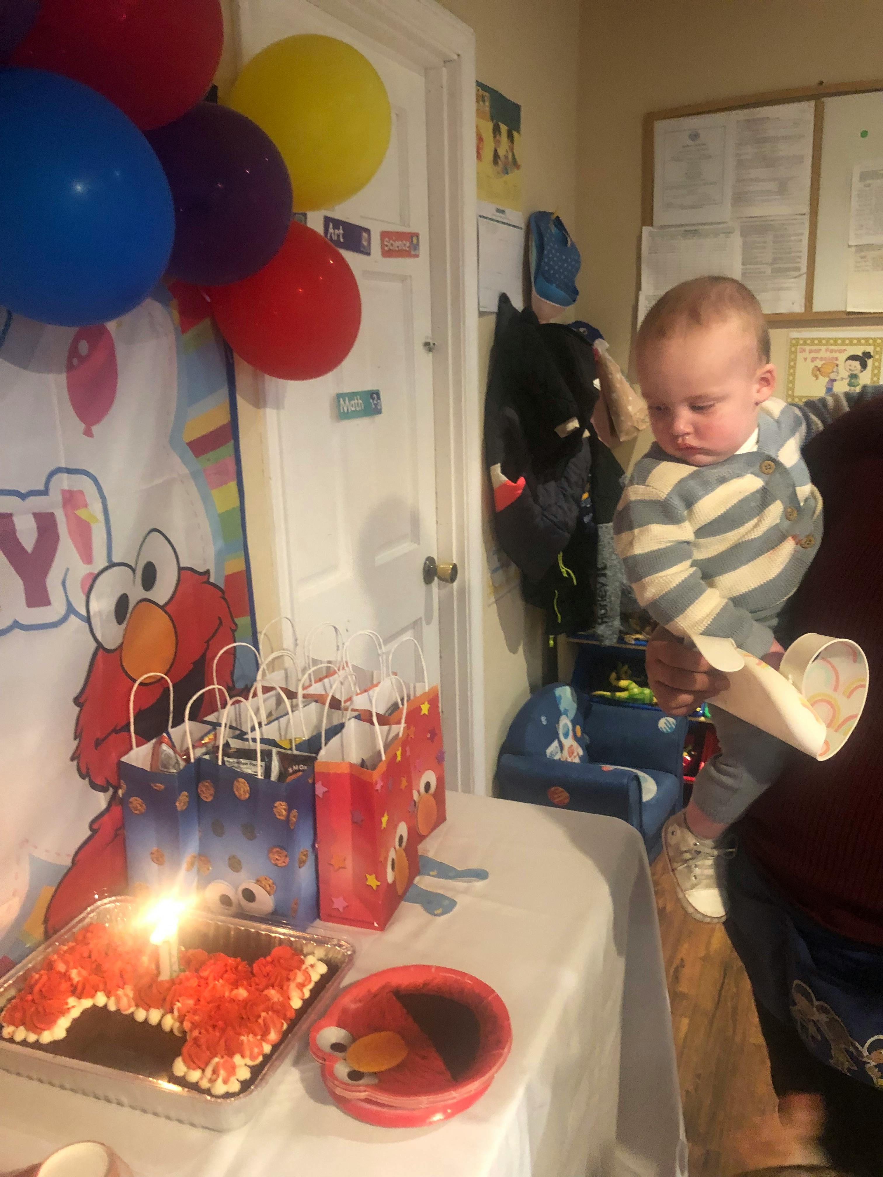 Child holding a cup near a table with a cake, gift bags, and balloons
