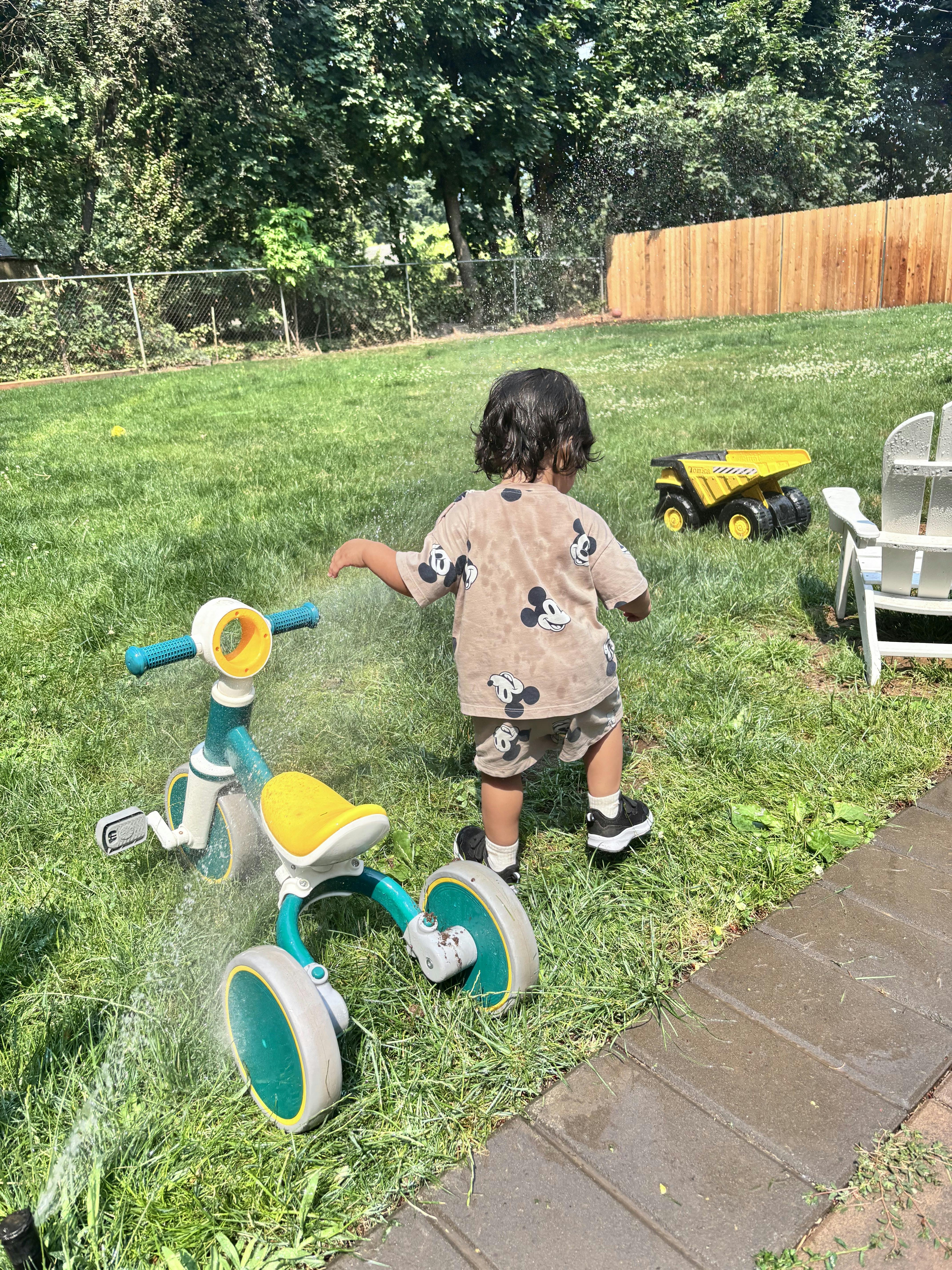 Child playing in a backyard with a tricycle and water sprinkler