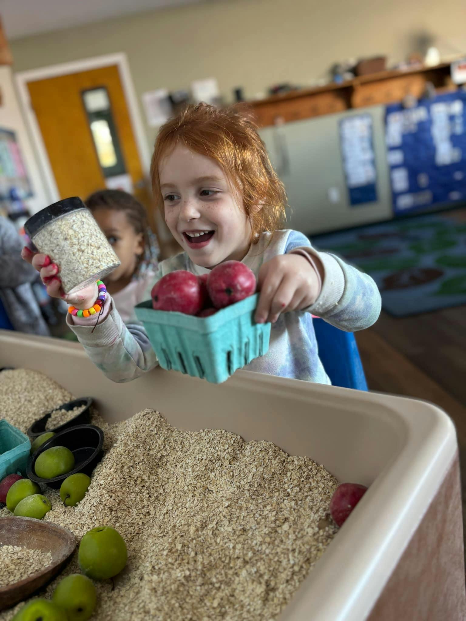 Child holding a container of oats and a basket of red apples over a sensory bin