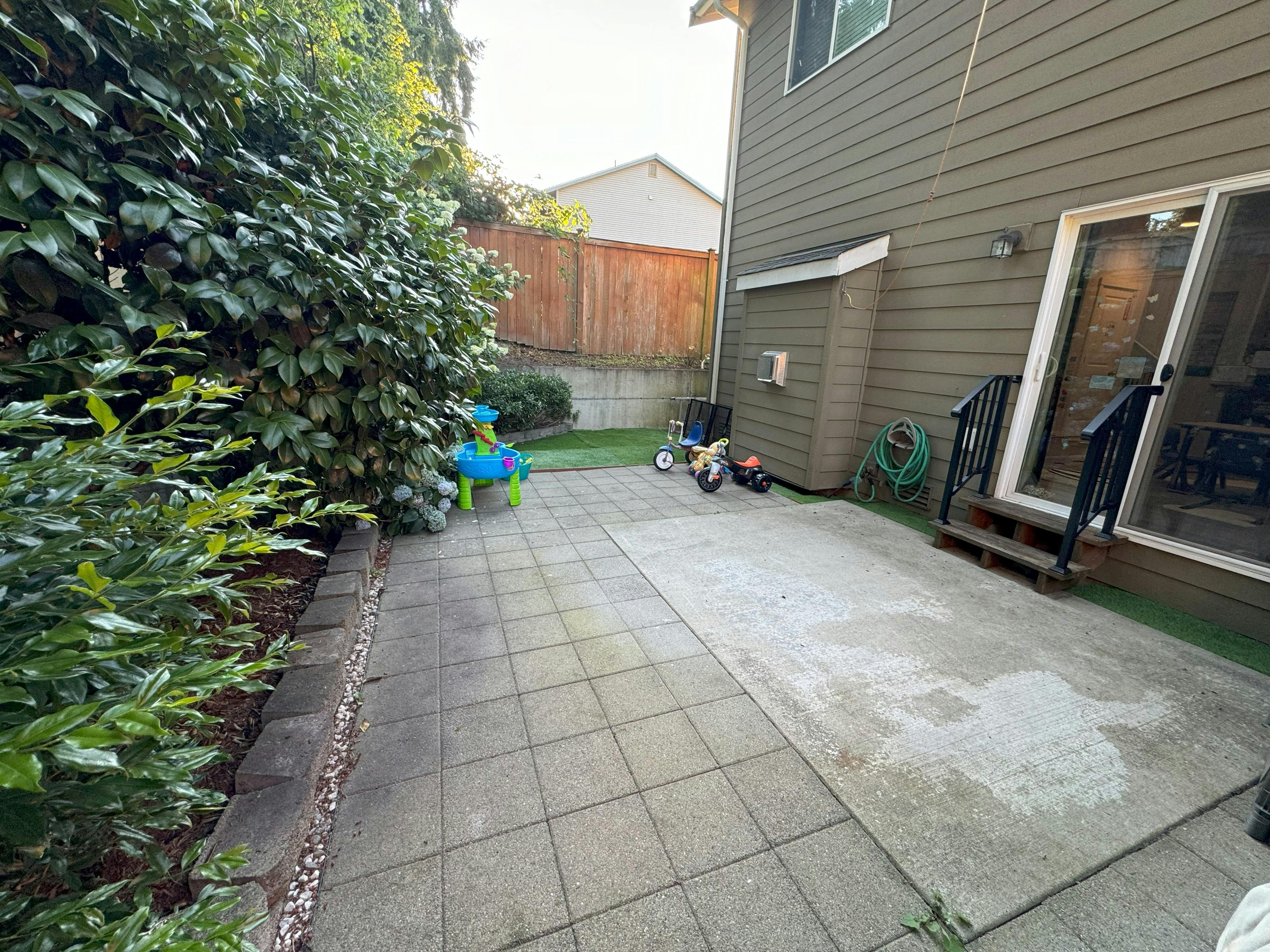 Backyard patio with a house, green bushes, and a wooden fence