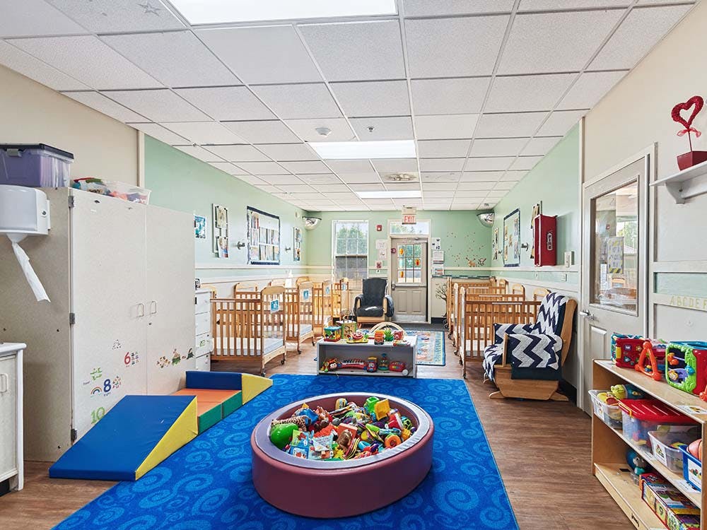 A childcare room with cribs, play mats, and toys on a blue patterned rug