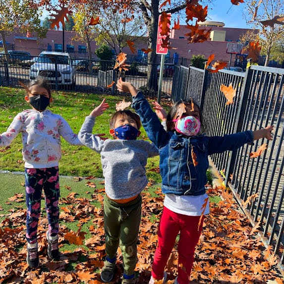 Three children in masks playing with autumn leaves