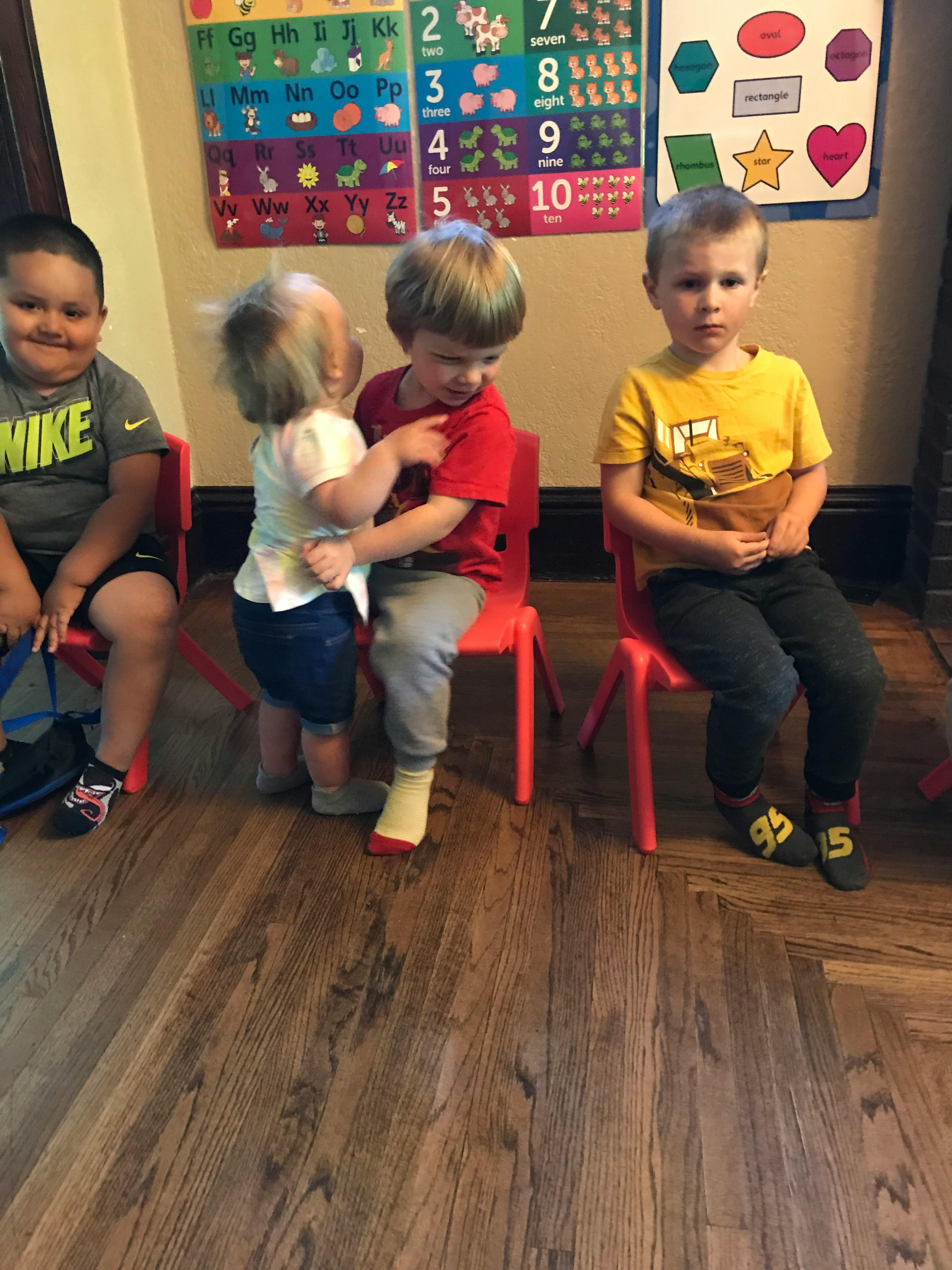 Three children sitting on small chairs, one child standing between two others