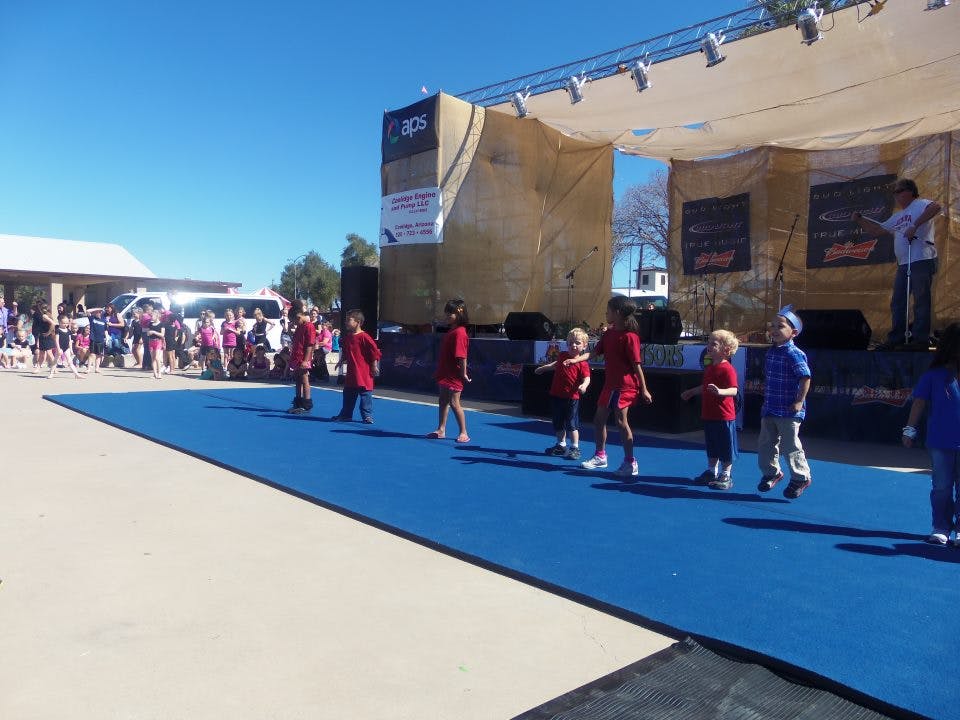 Children on a blue mat in front of a stage with an audience