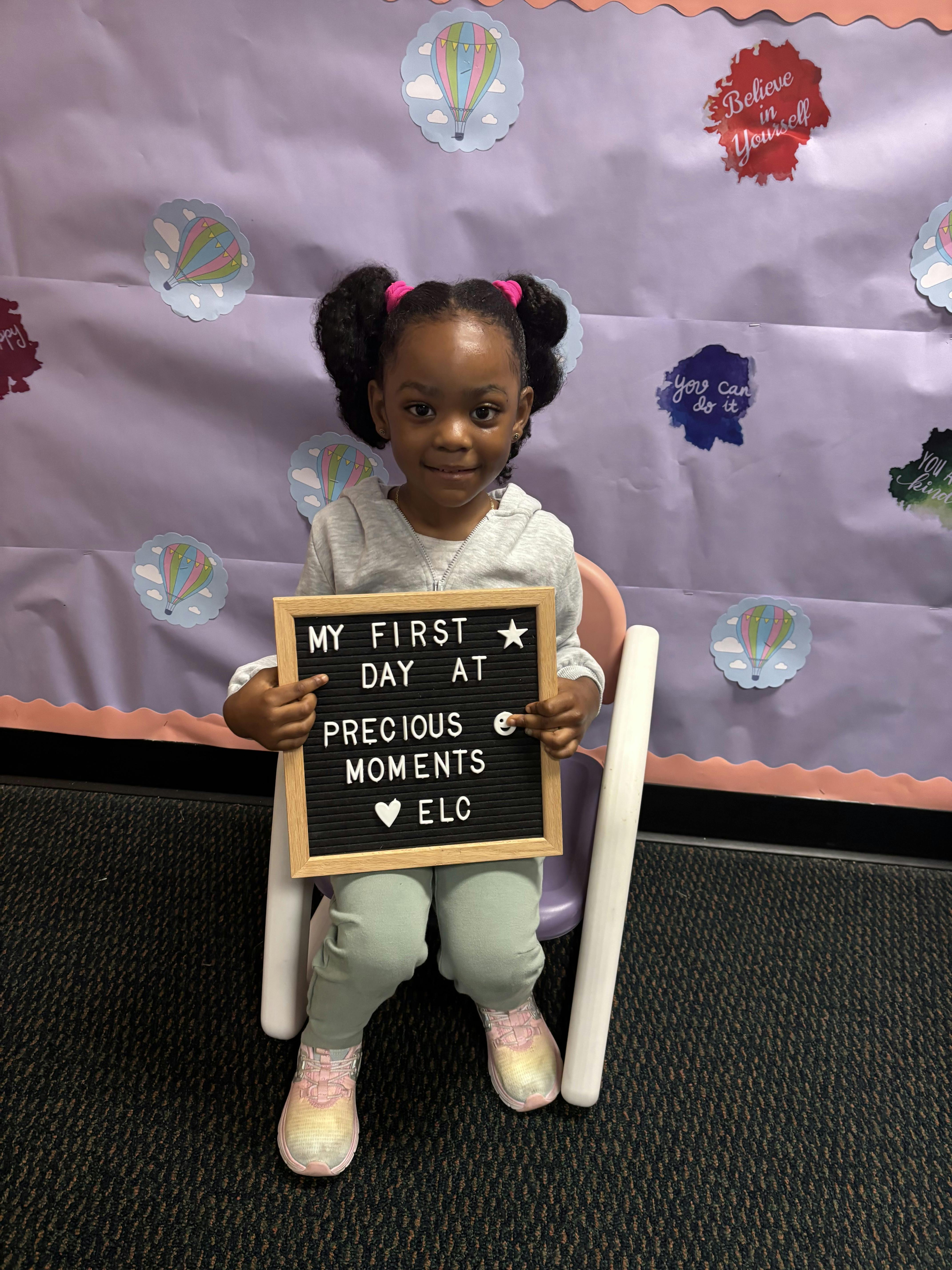 Child sitting in a chair holding a sign that reads "MY FIRST DAY AT PRECIOUS MOMENTS ELC"