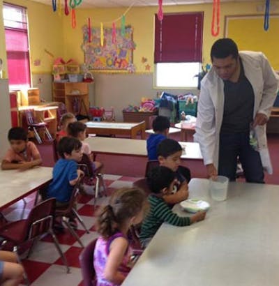 Person in lab coat with children at tables in a classroom