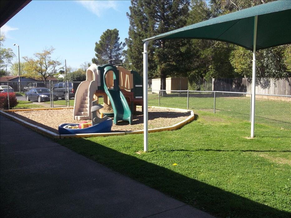 A playground with a slide and climbing structure on wood chips, next to a grassy area