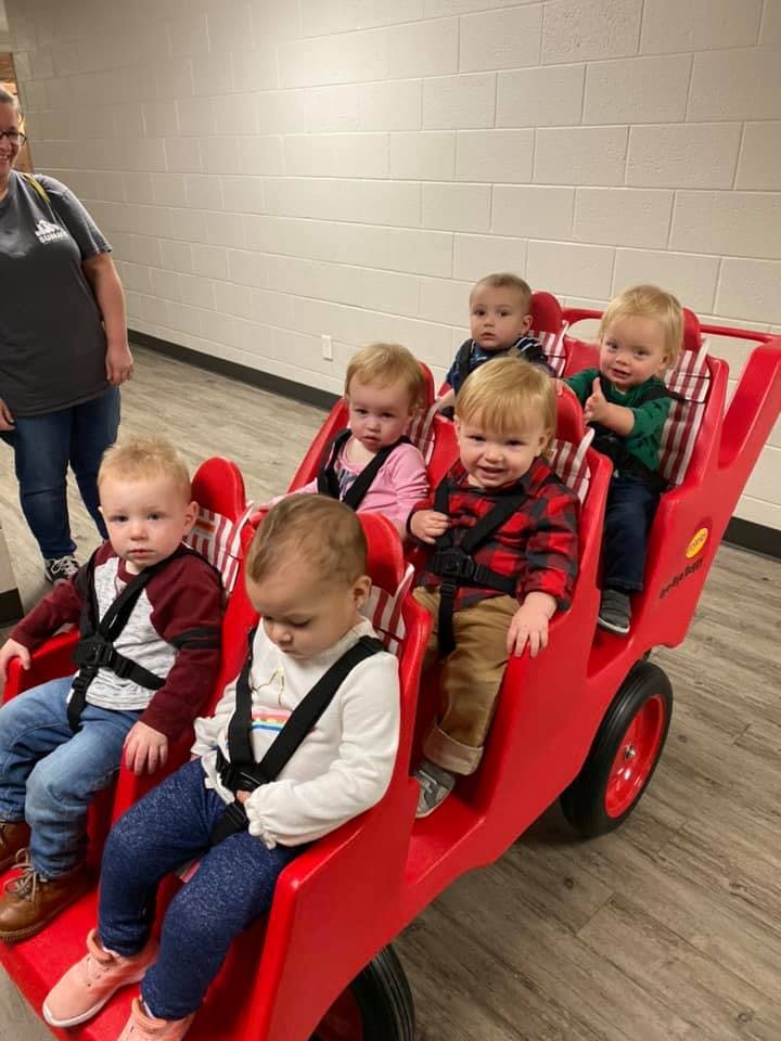 Six children in a red multi-seat stroller with a person standing nearby