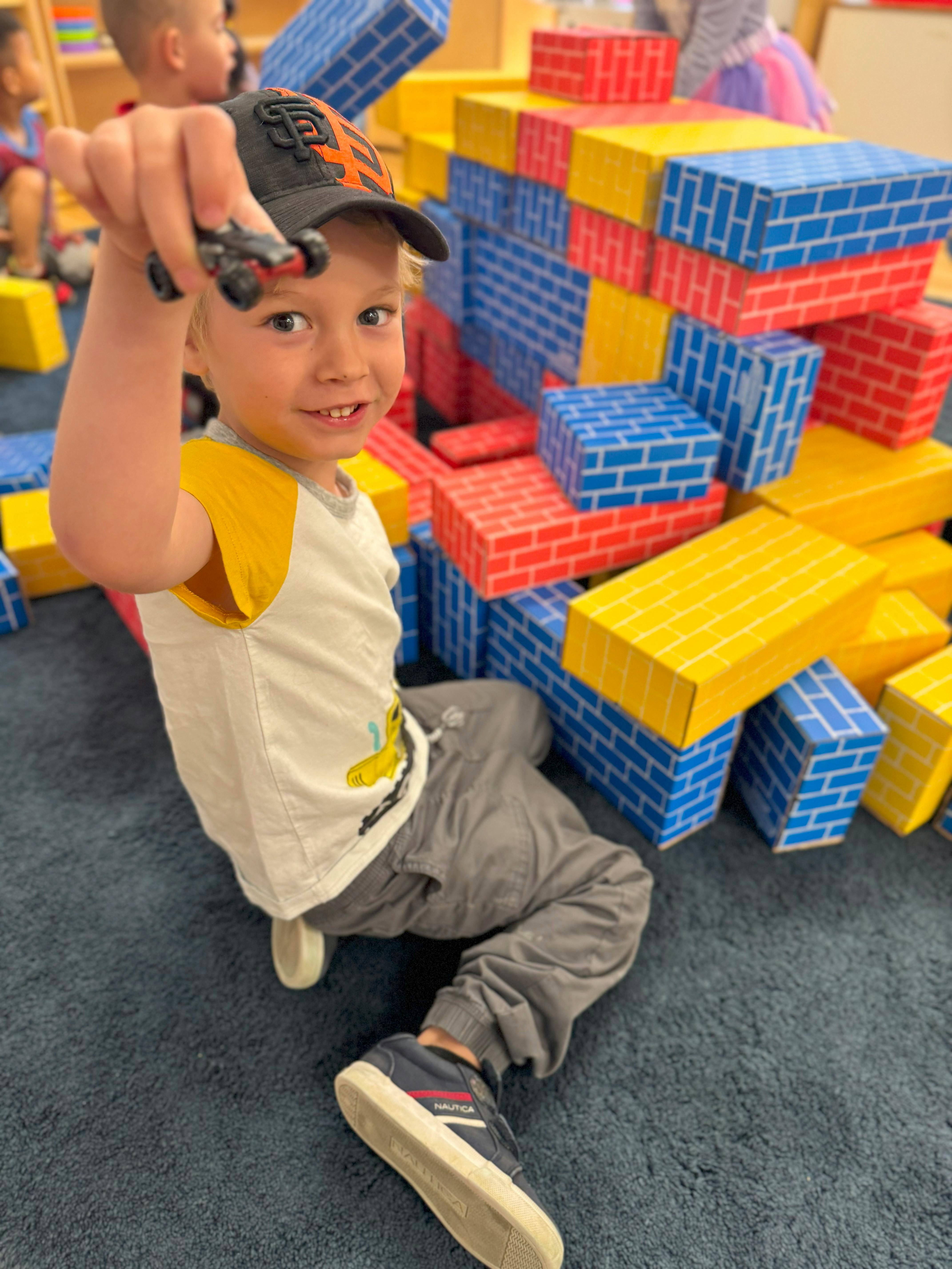 Child in a baseball cap holding up a toy car, surrounded by large building blocks