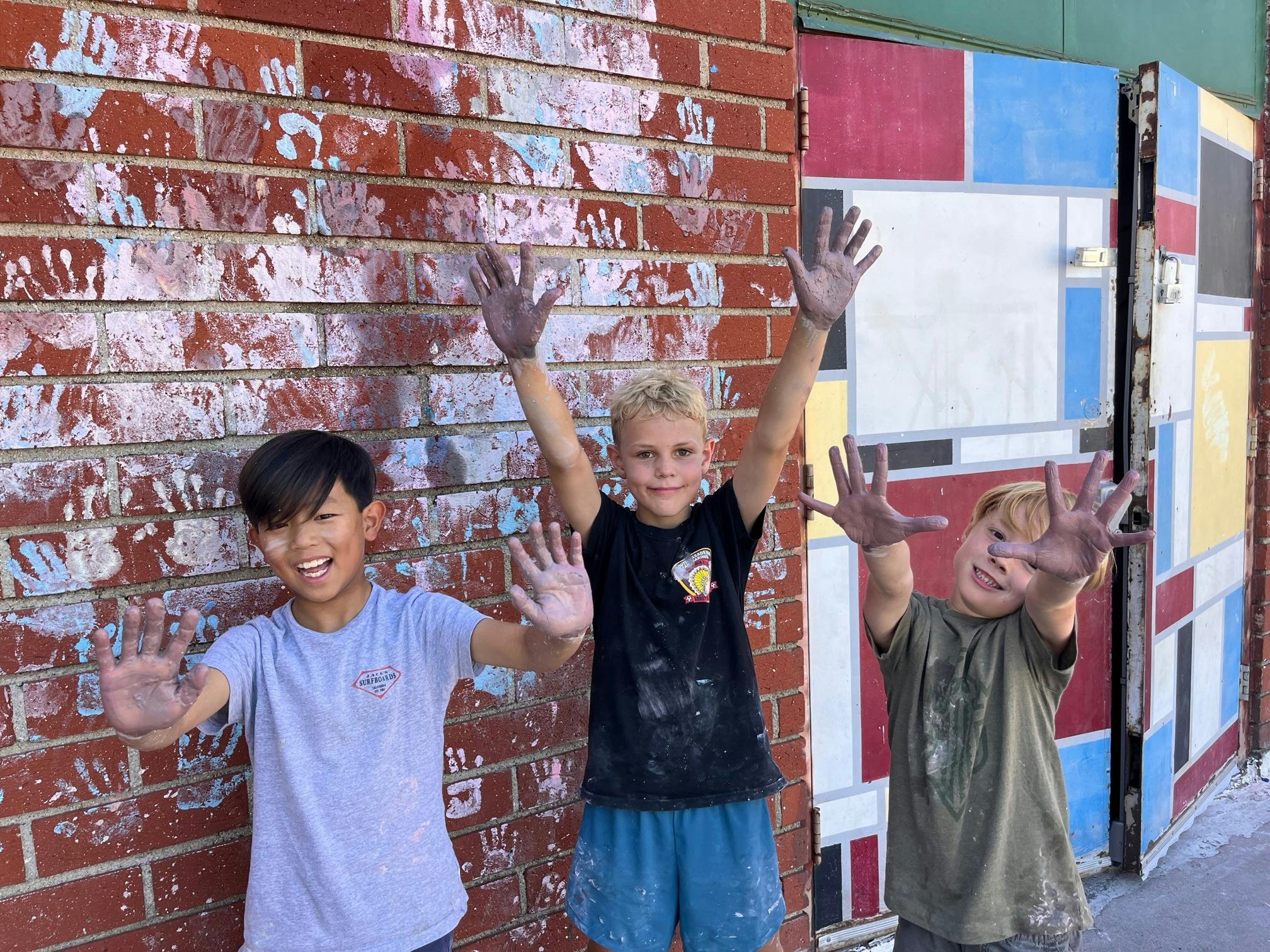Three children with paint on their hands standing in front of a brick wall