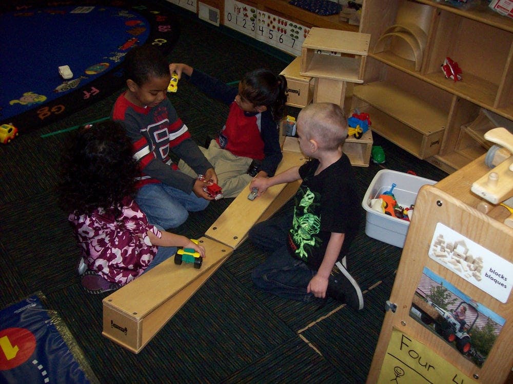 Four children playing with toy cars on wooden ramps in a classroom