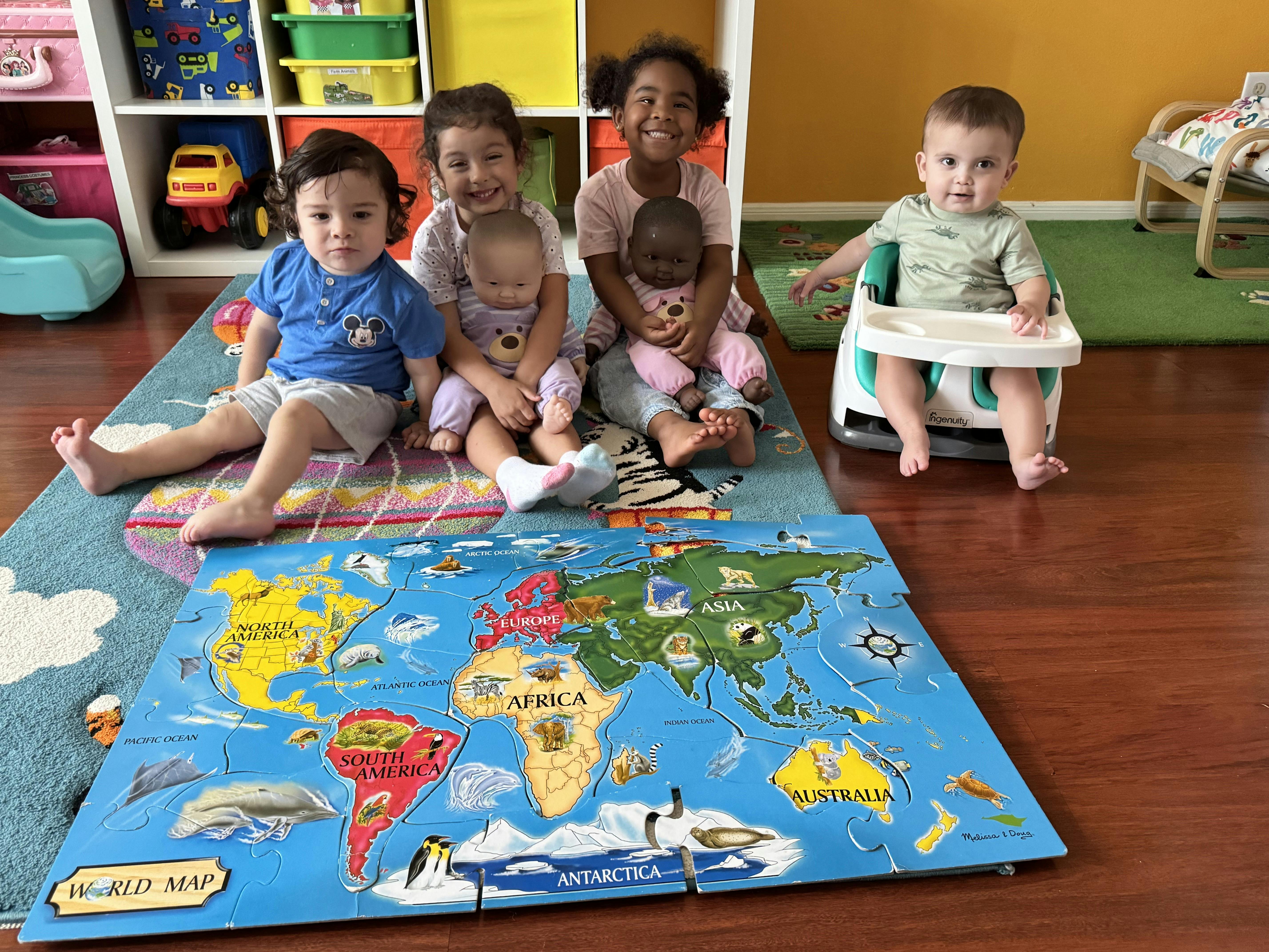 Four children and two baby dolls sitting on a rug with a world map puzzle
