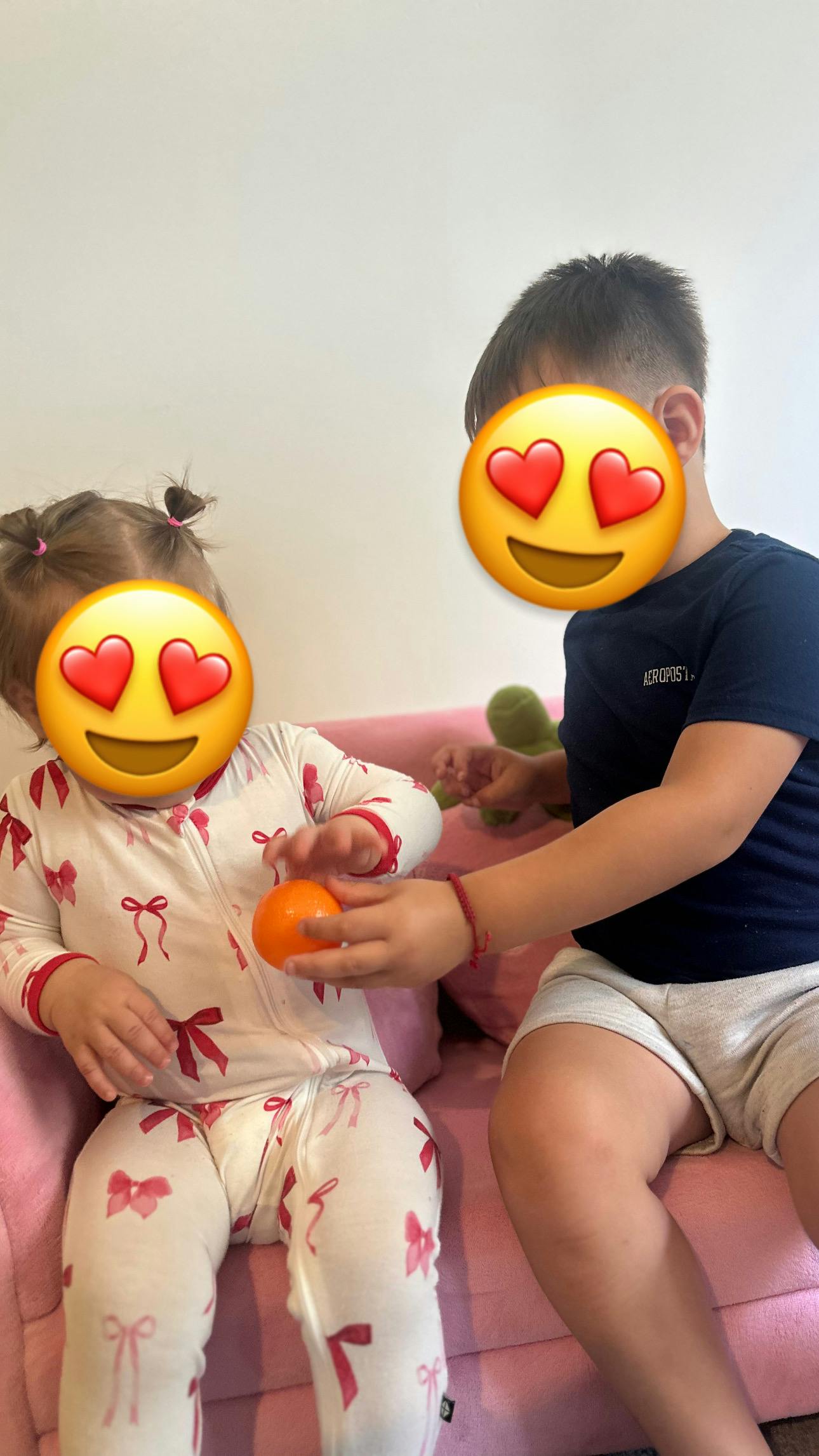 A teen and a child sitting on a pink couch while holding a small orange fruit