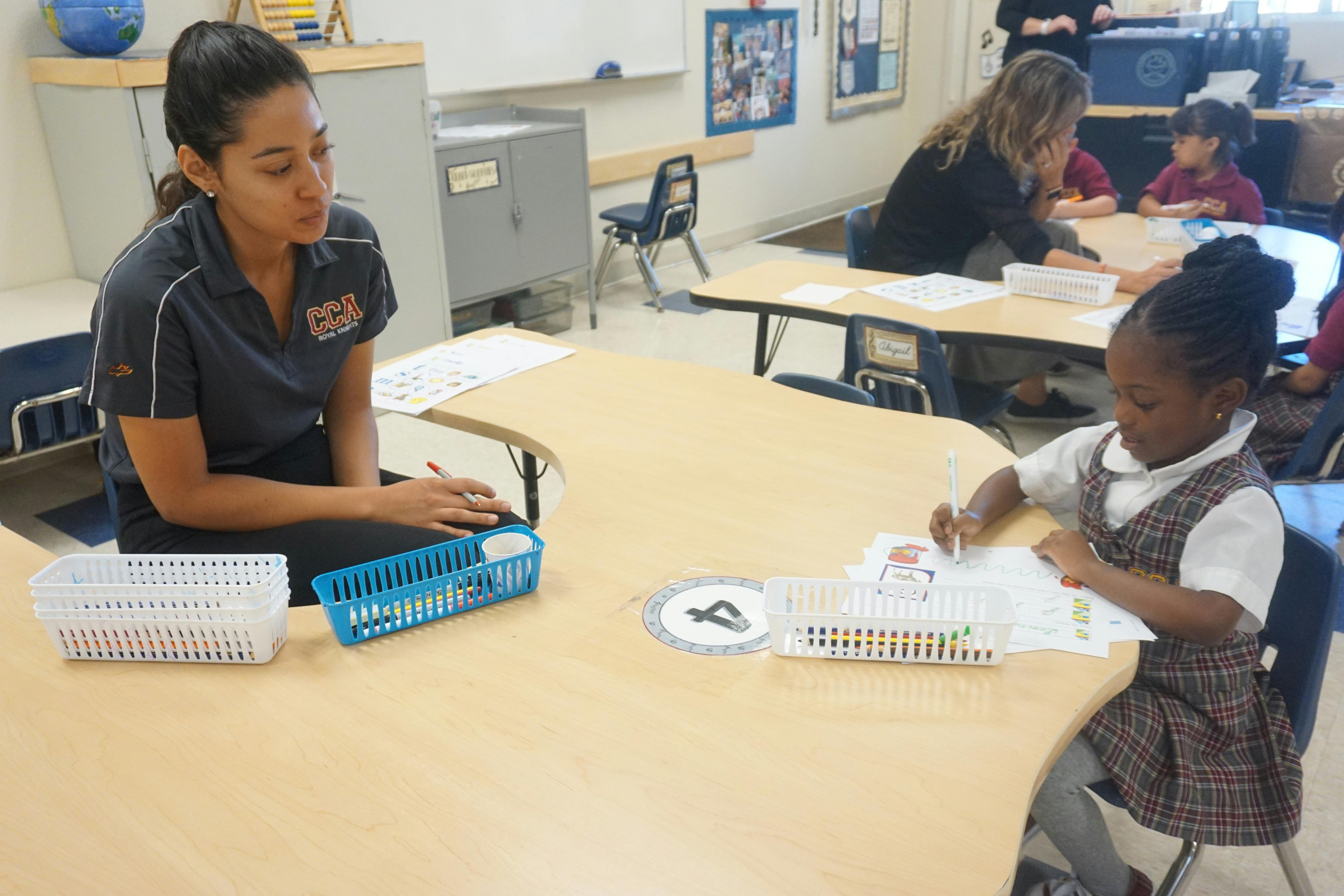 Person sitting at a desk with a child writing in a classroom
