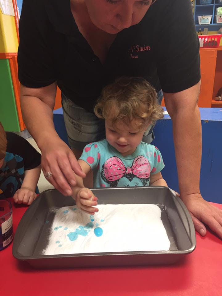 Person and child playing with blue drops in a tray of white material