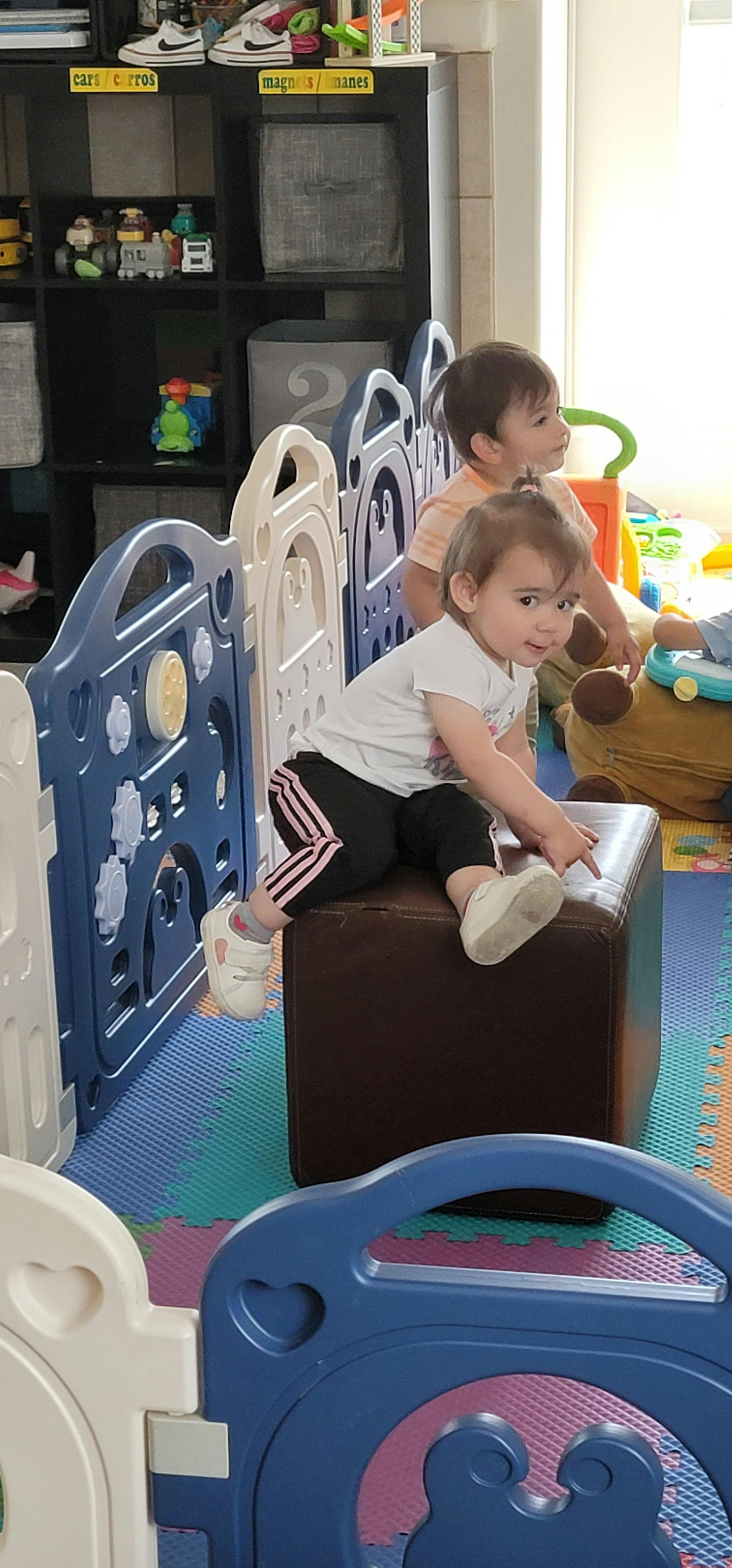 Child sitting on a brown ottoman, another child behind a playpen
