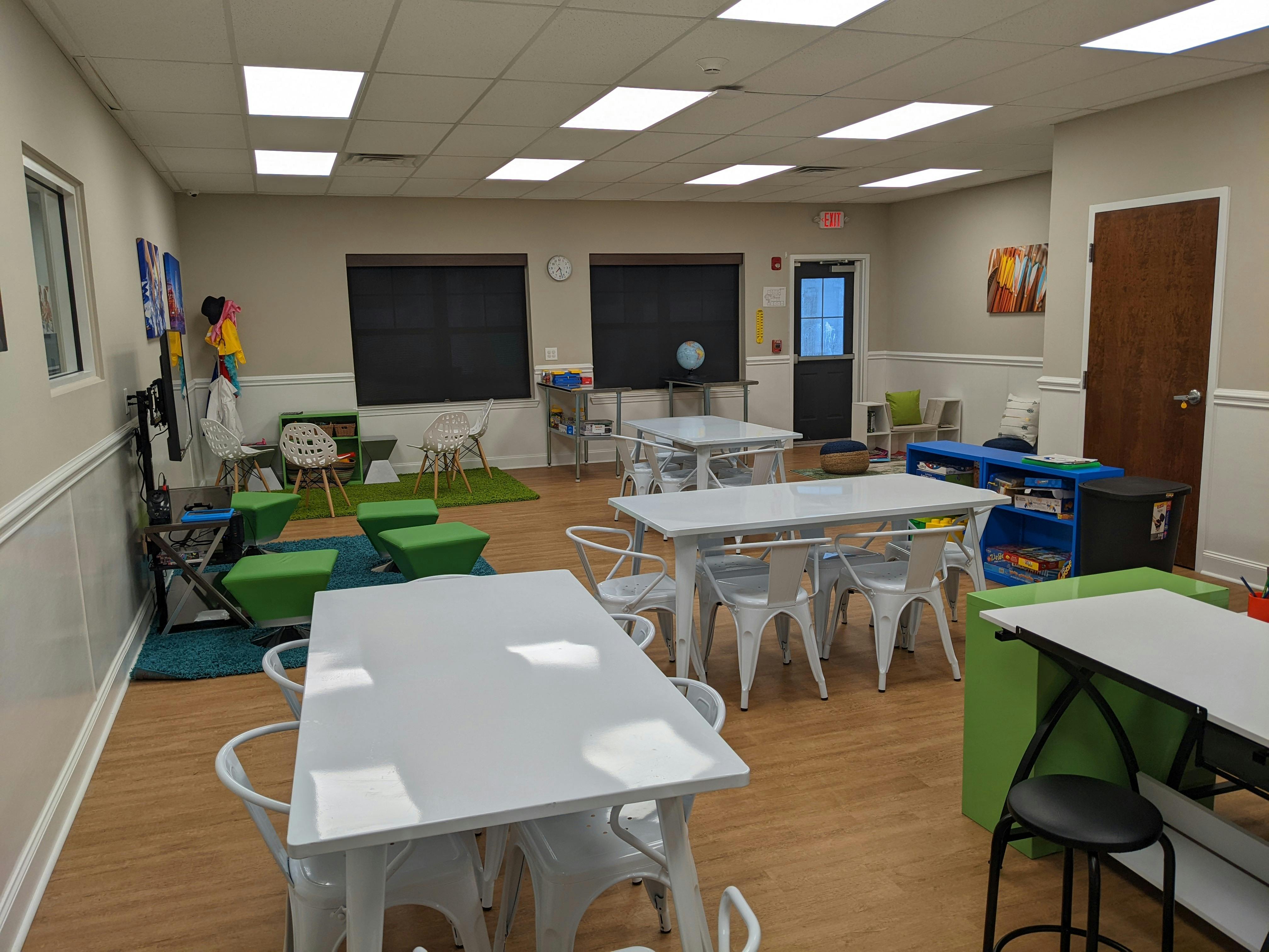 Classroom with white tables and chairs, green ottomans, and blackboards