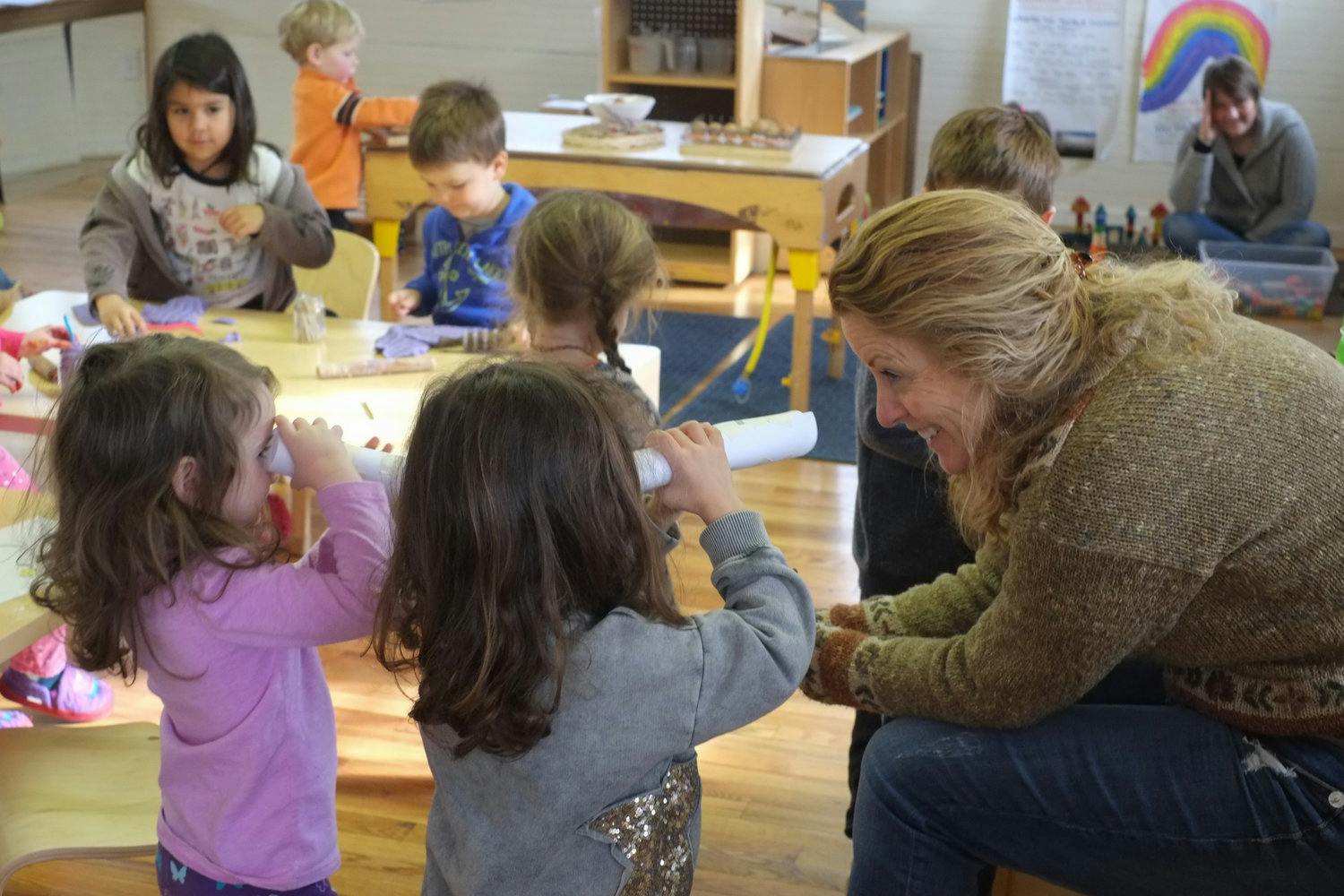 Adult and children in a classroom, two children holding paper tubes to their ears