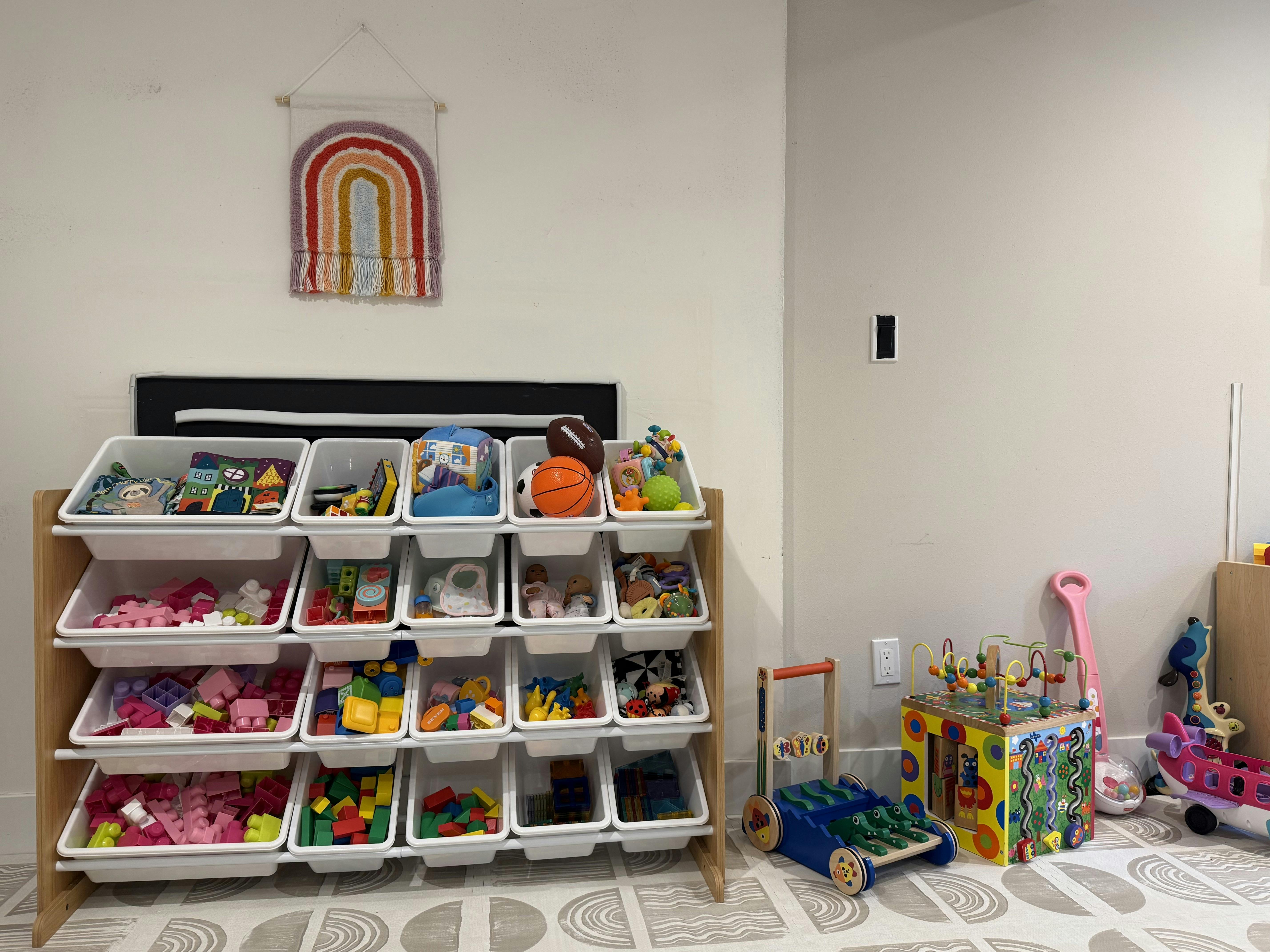 A wooden toy storage shelf filled with white bins of toys, and other toys on the floor