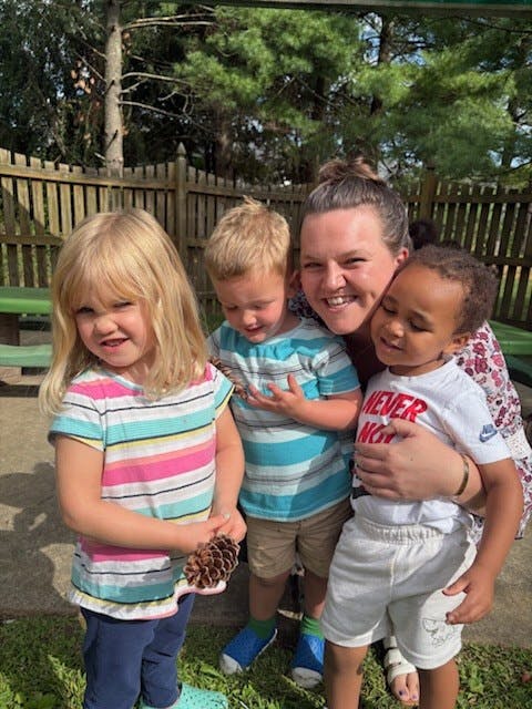 Smiling person with three young children outdoors, one child holds a pinecone