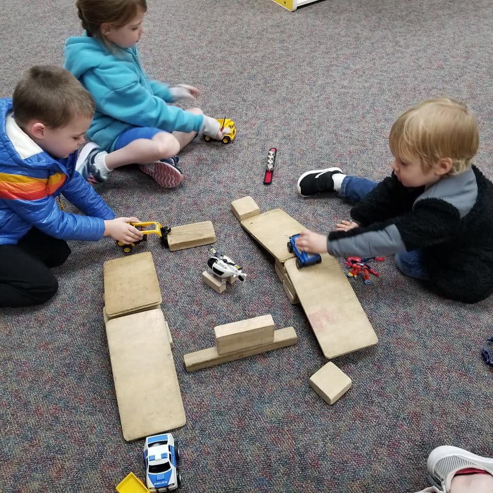 Three children playing with toy cars and wooden blocks on a carpeted floor