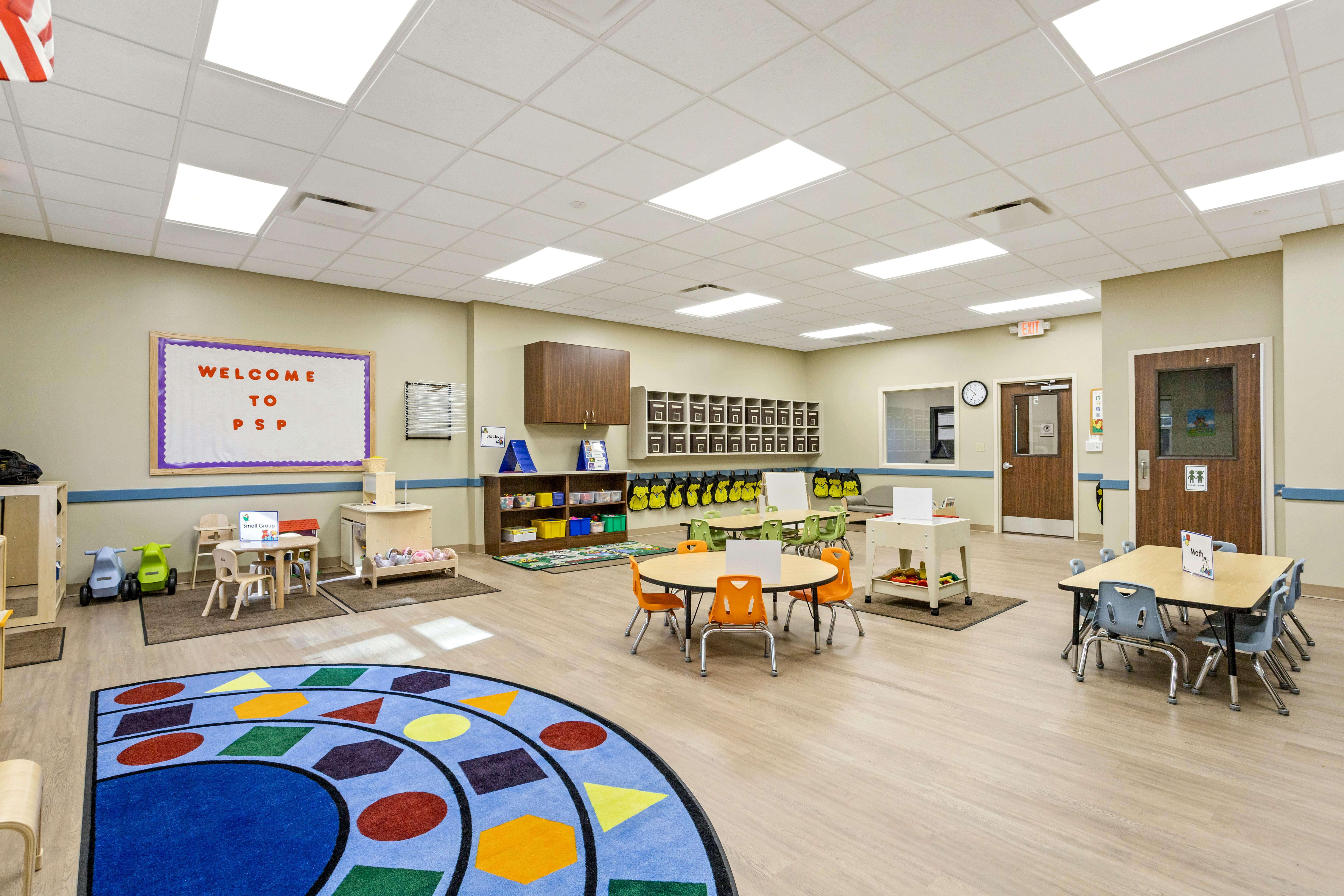 A colorful classroom with child-sized tables and chairs, and a large shape rug