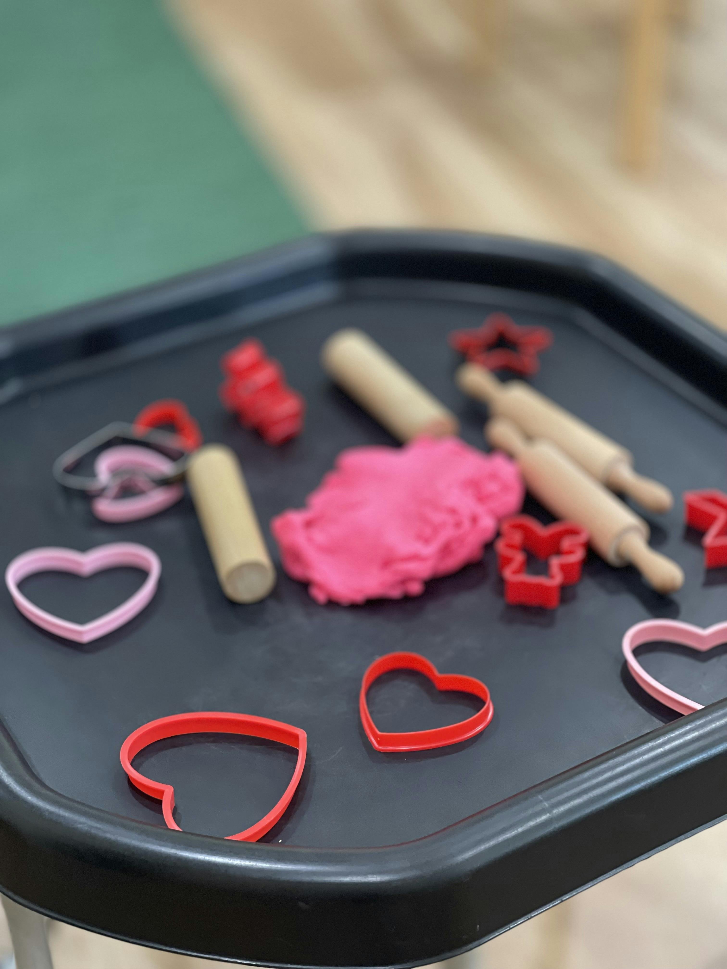 Pink playdough with cookie cutters and rolling pins on a black tray