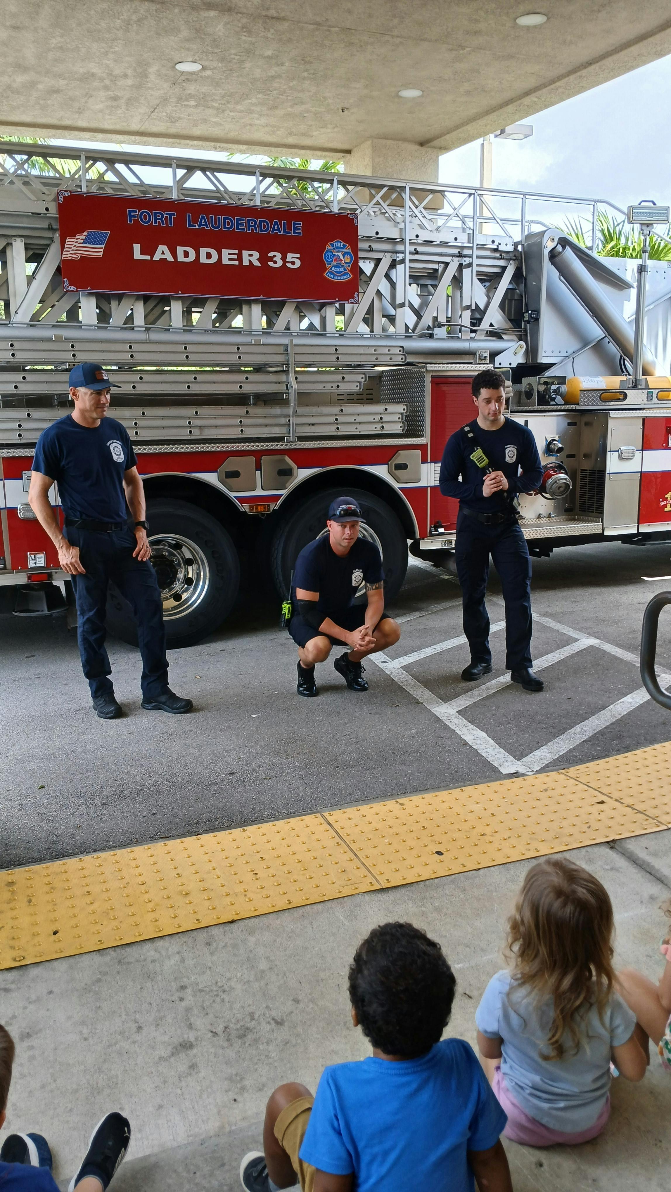 Three people in uniform standing and crouching near a fire truck, facing children