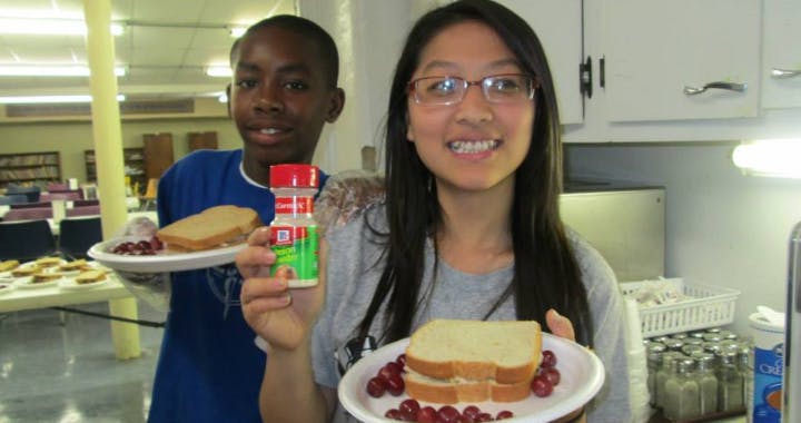 Two people holding plates with sandwiches and grapes, one holding a spice shaker