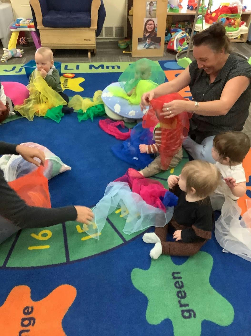 Adults playing with colorful sheer scarves with children on a classroom rug