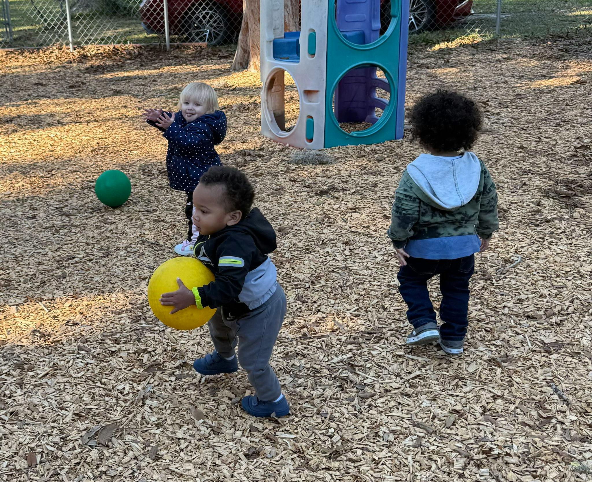 Three children playing with balls on a wood chip surface