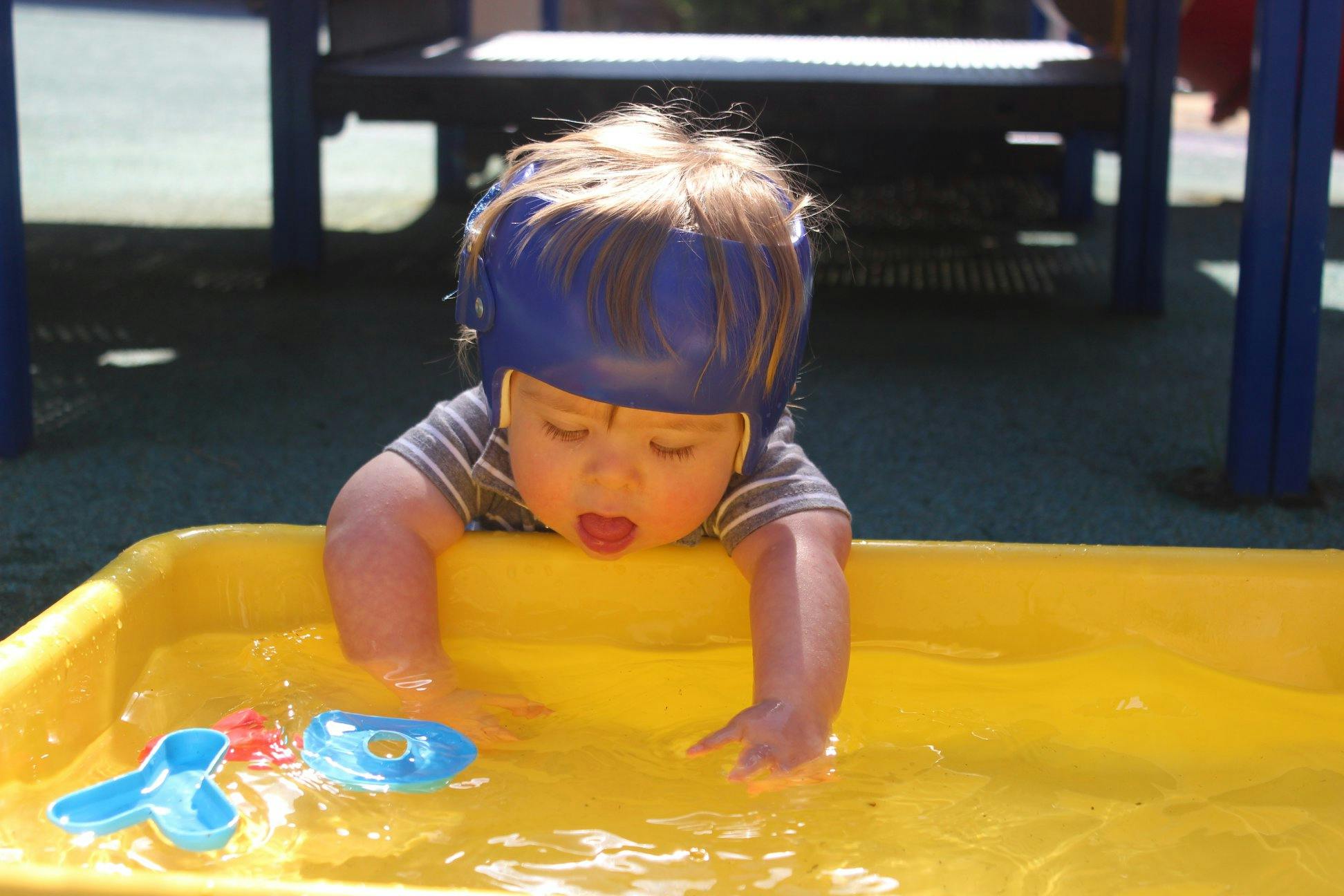 Child in a blue helmet playing with water in a yellow bin